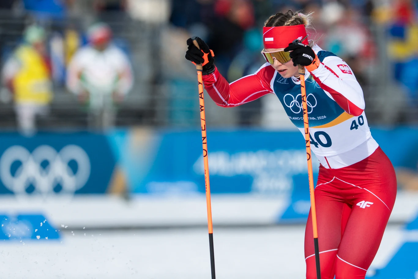 Zawodniczka w czerwono-białym stroju sportowym podczas rywalizacji w biegach narciarskich na zimowych igrzyskach olimpijskich, w tle widoczne rozmazane sylwetki oraz logo olimpijskie.