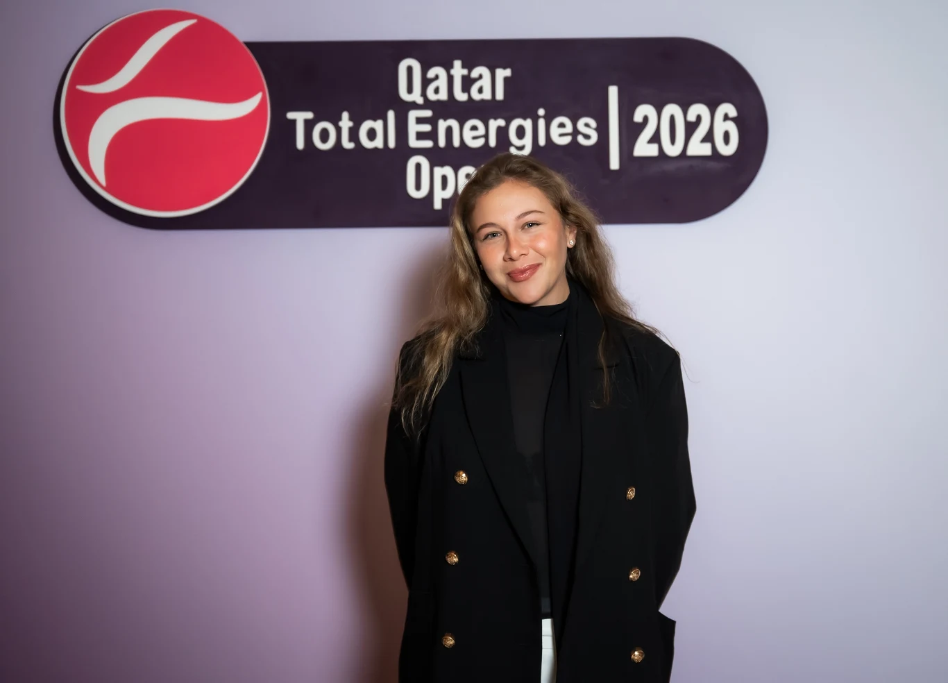 Amanda Anisimova Kobieta w czarnej marynarce i ciemnym golfie stoi na tle tablicy z logo Qatar Total Energies Open 2026, delikatnie uśmiechając się do aparatu.