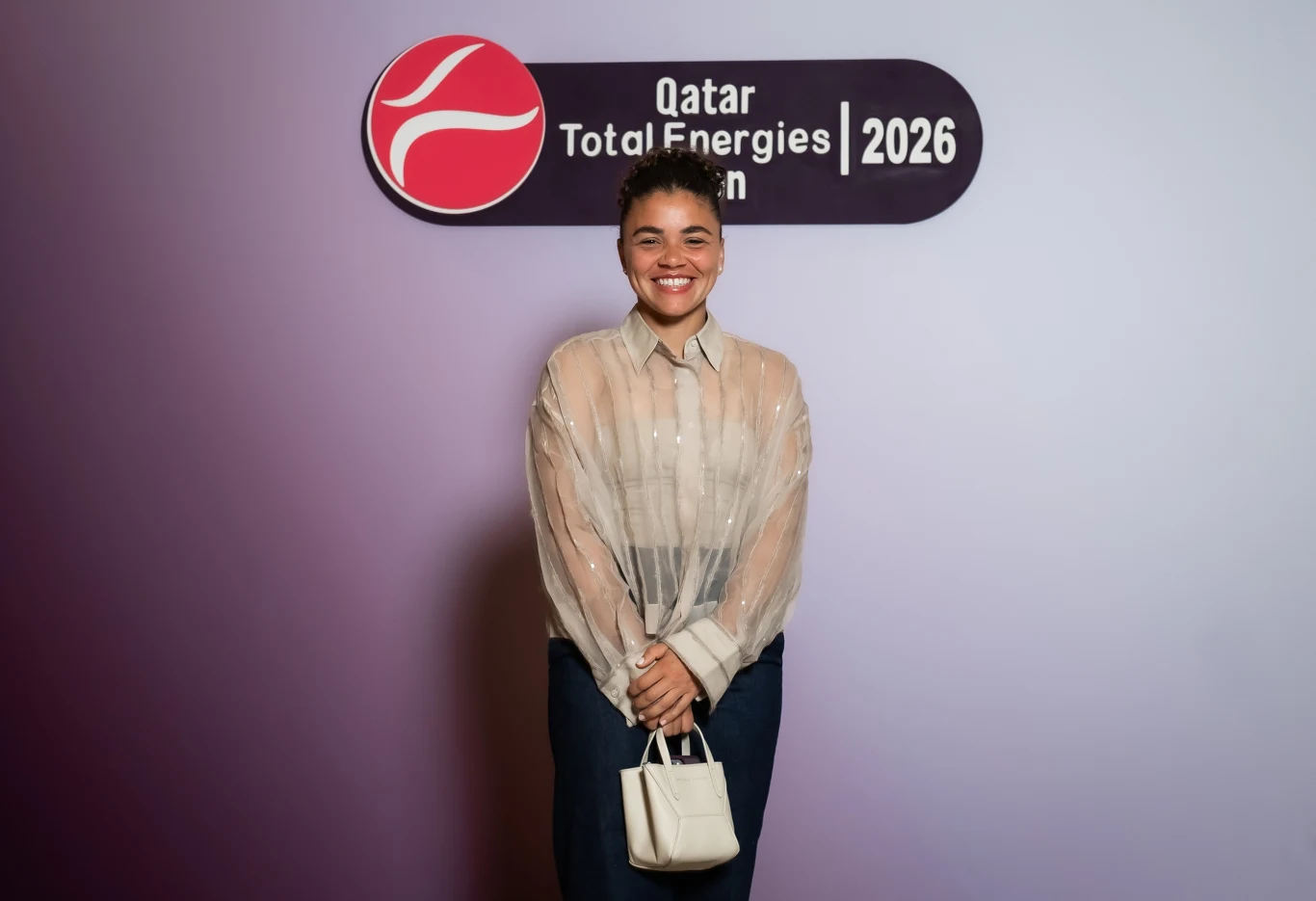 Jasmine Paolini Uśmiechnięta kobieta stojąca na tle ścianki z logo i napisem Qatar Total Energies Open 2026, ubrana w jasną bluzkę i trzymająca białą torebkę.
