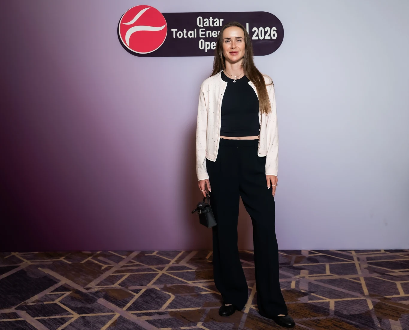 Elina Svitolina Kobieta w eleganckim, czarnym ubraniu i jasnej marynarce pozuje na tle planszy z napisem Qatar Total Energies Open 2026. Trzyma w dłoni torebkę, stoi na wykładzinie o geometrycznym wzorze.