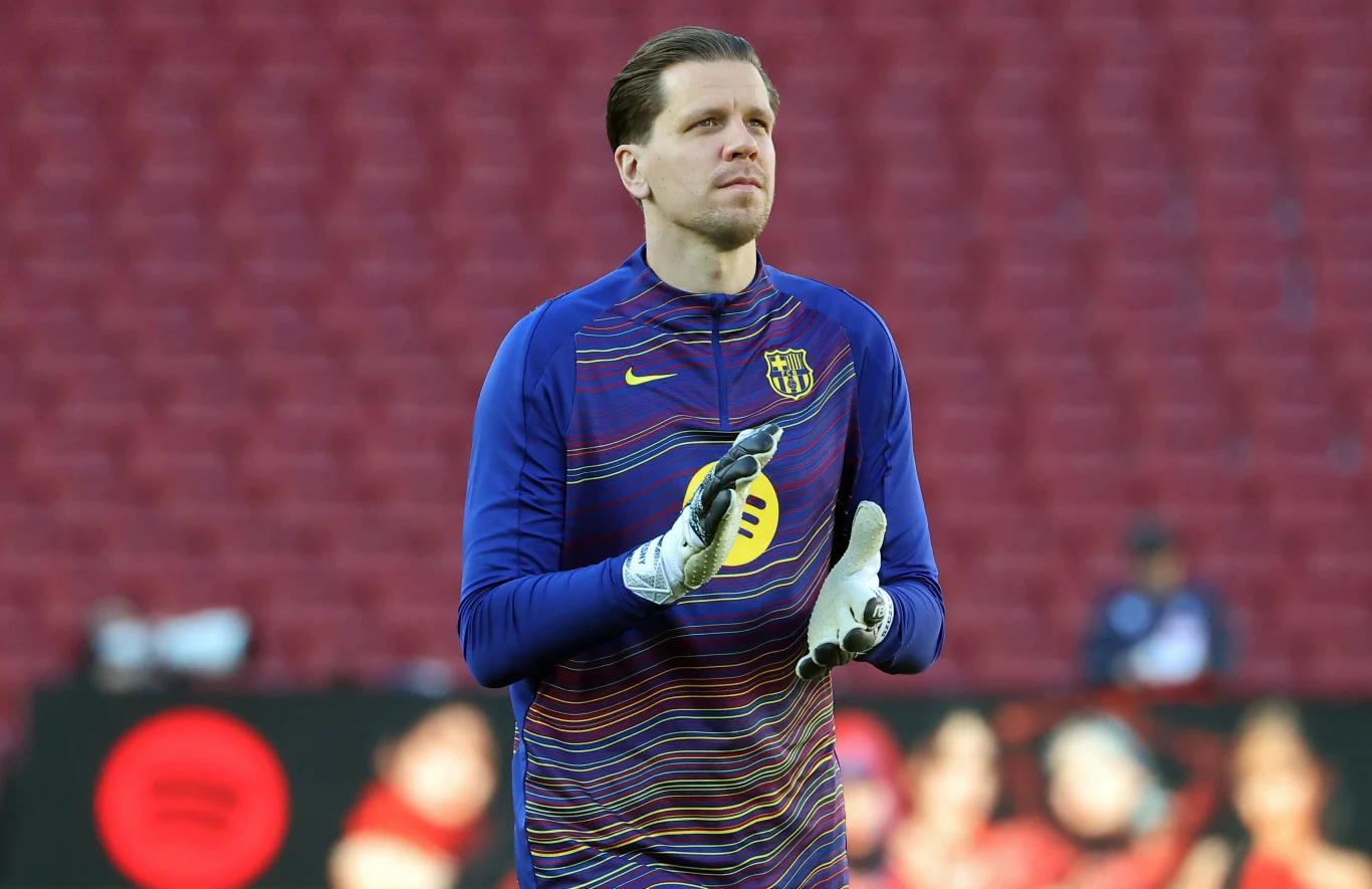 Wojciech Szczęsny Piłkarz w sportowej bluzie z rękawicami bramkarskimi stoi na tle pustych trybun stadionu, patrząc przed siebie i sprawiając wrażenie skoncentrowanego.