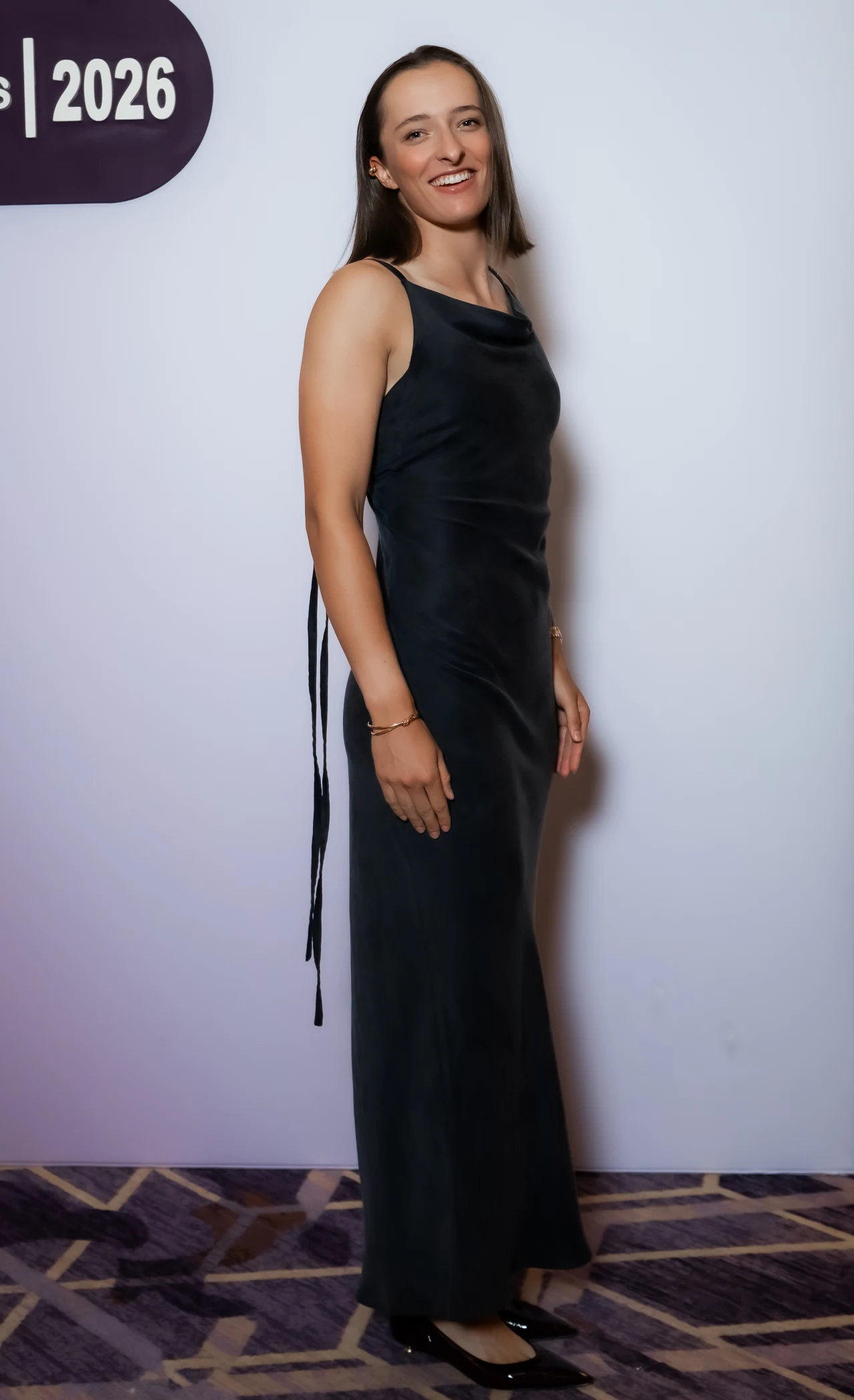 Iga Świątek Kobieta w długiej, ciemnej, eleganckiej sukni stoi bokiem na tle jasnej ściany z fioletowym napisem i liczbą 2026. Ma rozpuszczone włosy, delikatną biżuterię oraz czarne buty na obcasie. Uśmiecha się i patrzy w kierunku aparatu.
