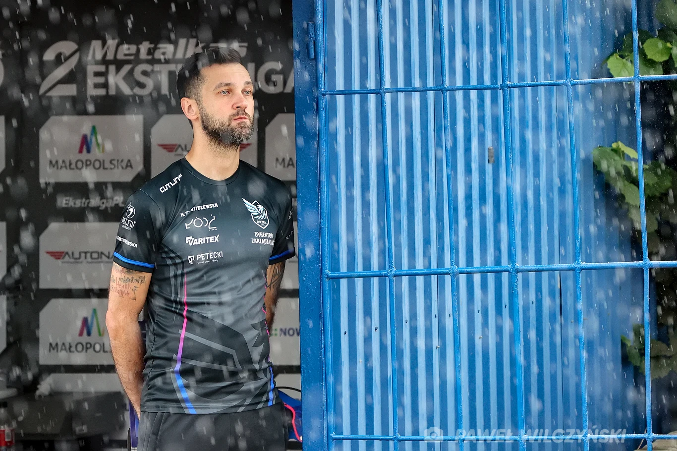 Mężczyzna w sportowej koszulce drużyny żużlowej stoi obok niebieskiej bramy, otoczony padającym śniegiem. W tle widoczne są tablice sponsorów i logotypy związane z wydarzeniem sportowym.