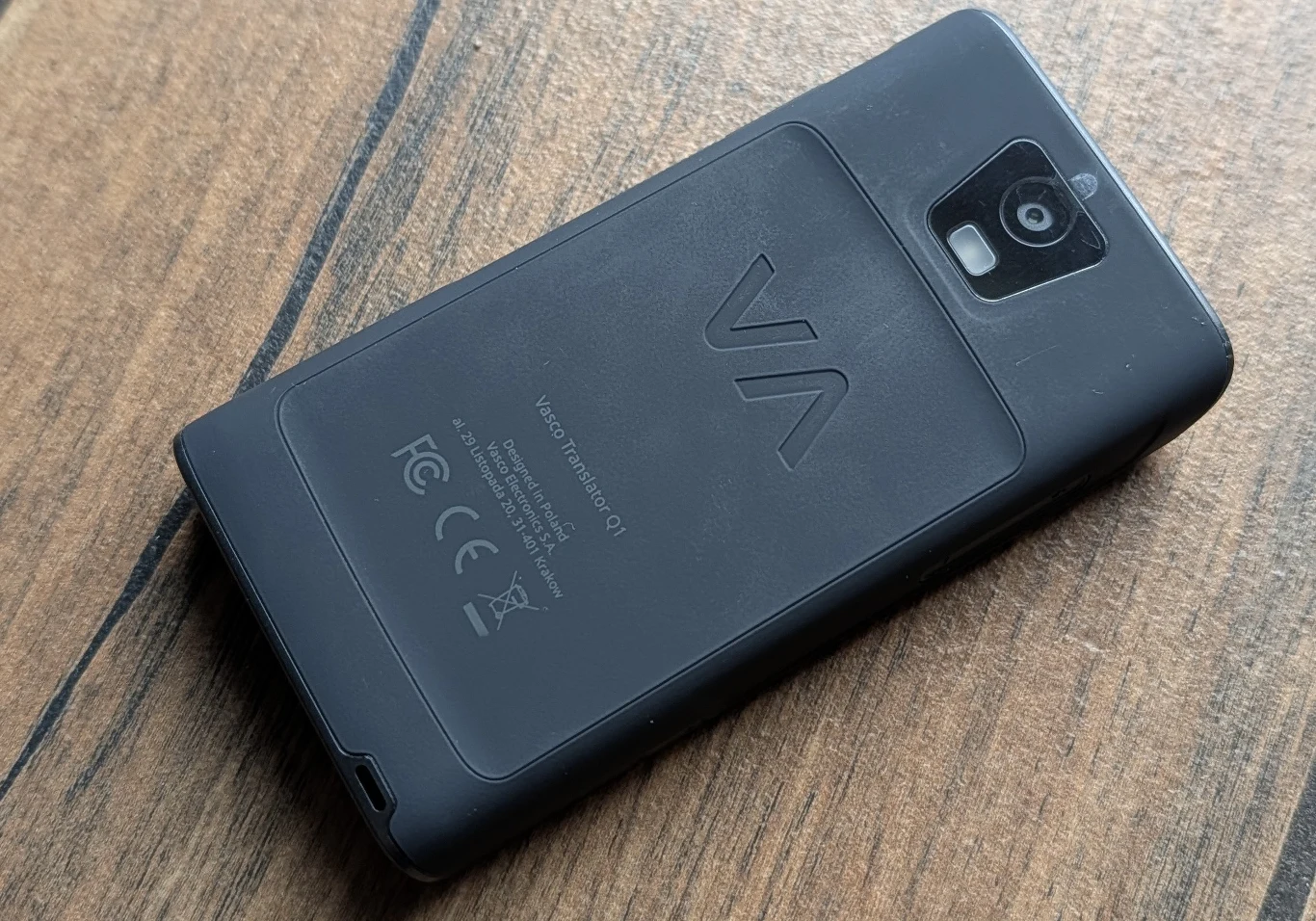 Czarny telefon komórkowy z widocznym aparatem i napisem na tylnej klapce, położony na drewnianym blacie.