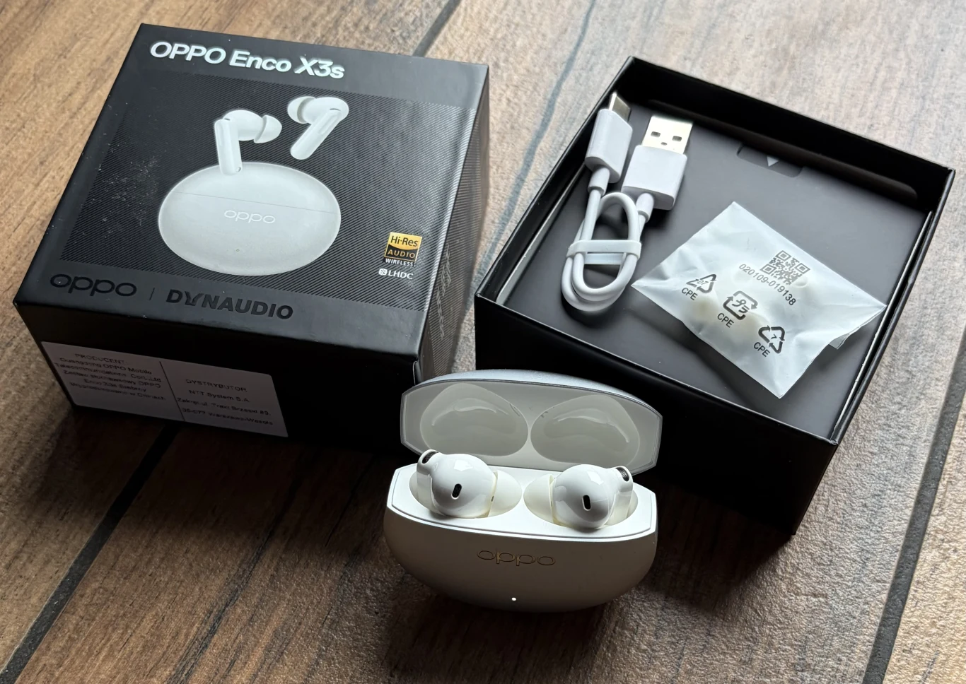 Zawartość opakowania OPPO Enco X3s. Bezprzewodowe słuchawki Oppo Enco X3s w białym etui ładującym ustawione obok oryginalnego opakowania, kabla USB-C oraz zapasowych nakładek, całość na drewnianym tle.