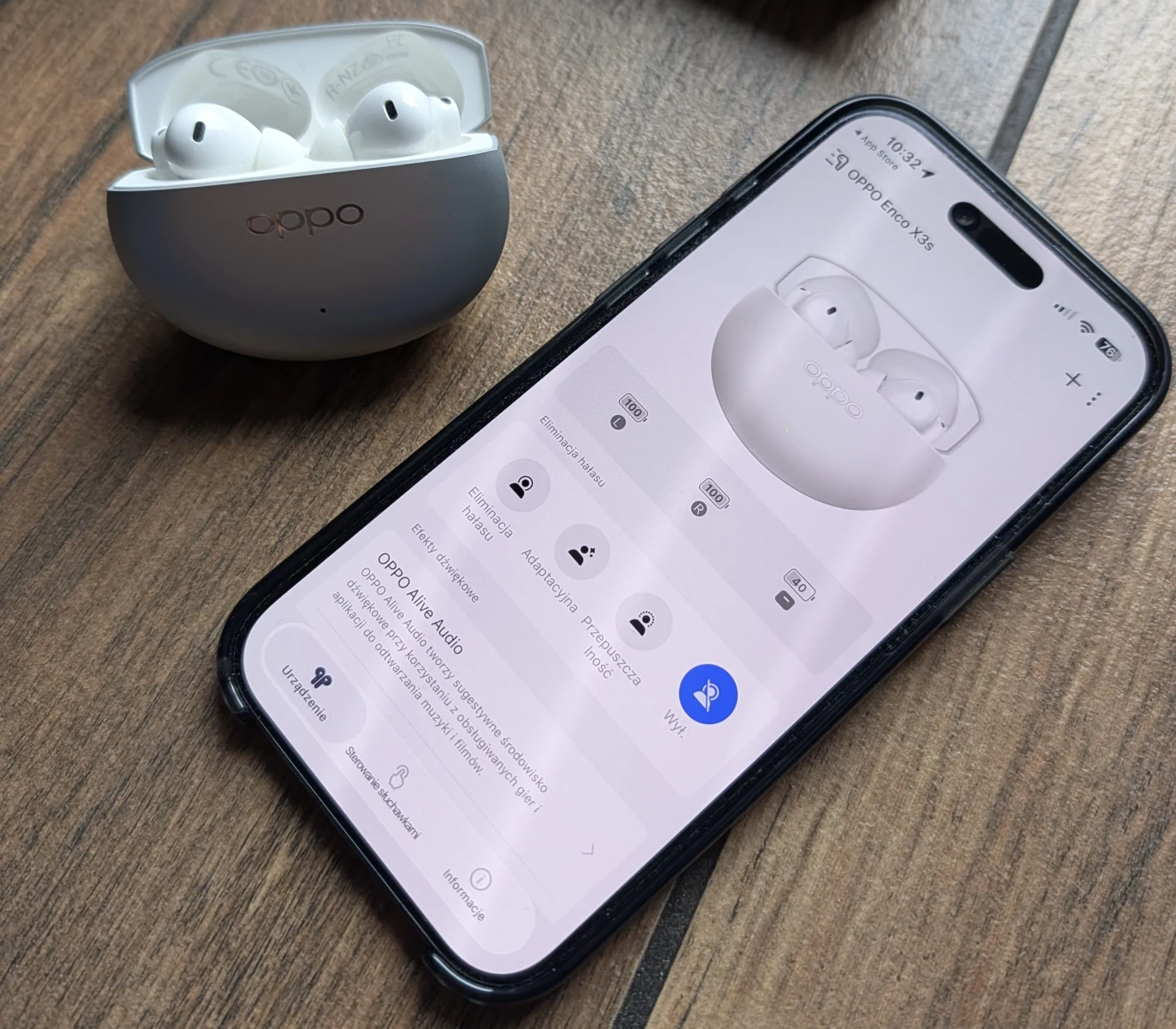 Słuchawki bezprzewodowe OPPO Enco X3s oferują różne tryby pracy. Etui z bezprzewodowymi słuchawkami Oppo leży obok smartfona z włączoną aplikacją do zarządzania tymi słuchawkami na drewnianym blacie, wyświetlacz telefonu pokazuje ustawienia dźwięku oraz opcje połączenia.