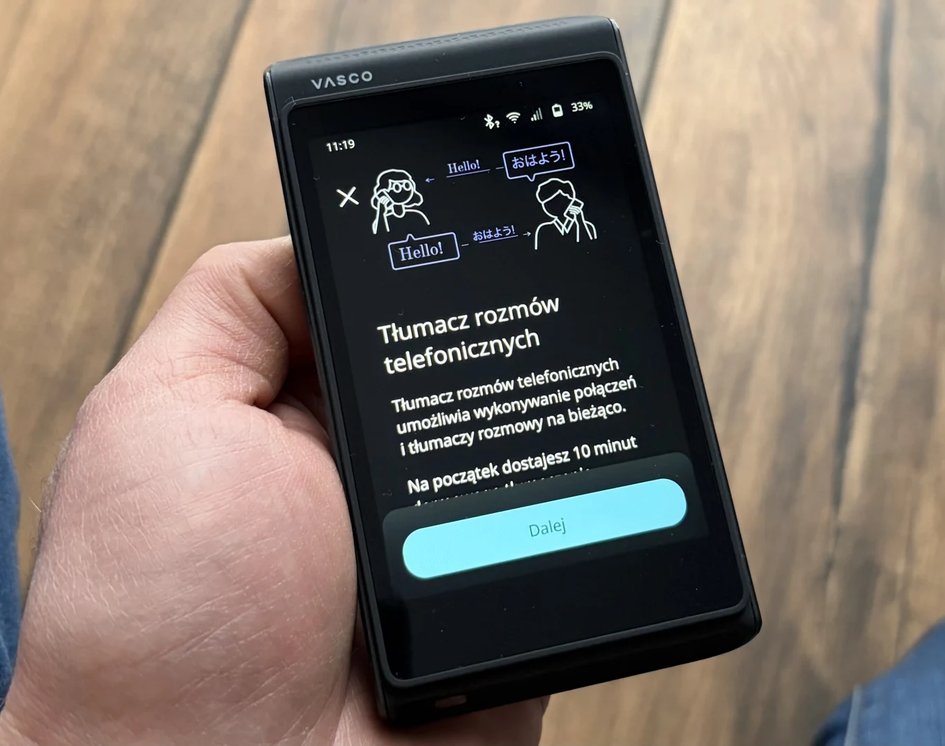 Ręka trzymająca urządzenie elektroniczne Vasco, na wyświetlaczu panel tłumacza rozmów telefonicznych z ilustracjami dwóch osób oraz informacją o dostępnych minutach na rozmowy.