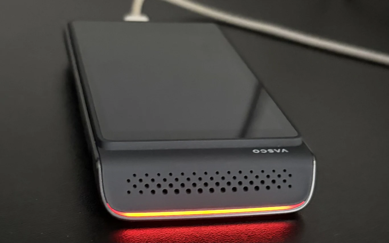 Czarny, kompaktowy powerbank marki VASCO leżący na ciemnej powierzchni, z podświetleniem LED w kolorze czerwonym widocznym na jednym z boków, oraz kablem podłączonym do urządzenia.