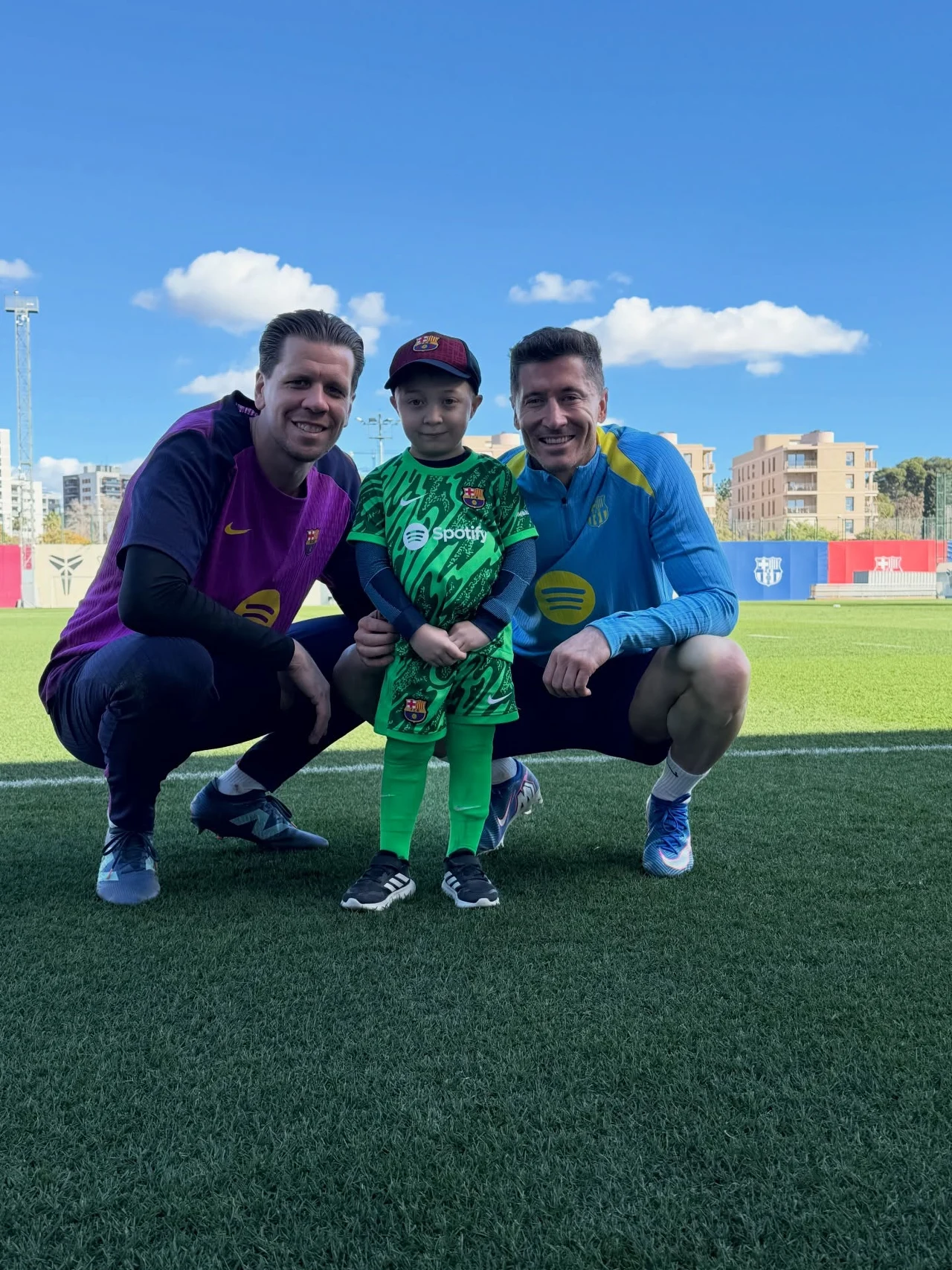 "Nieduży Wojtek" pozujący z Wojciechem Szczęsnym oraz Robertem Lewandowskim Dwóch dorosłych mężczyzn w sportowych strojach oraz mały chłopiec w zielonym stroju piłkarskim stoją razem na boisku piłkarskim, wszyscy uśmiechnięci, w tle widoczne budynki oraz niebieskie niebo z białymi chmurami.