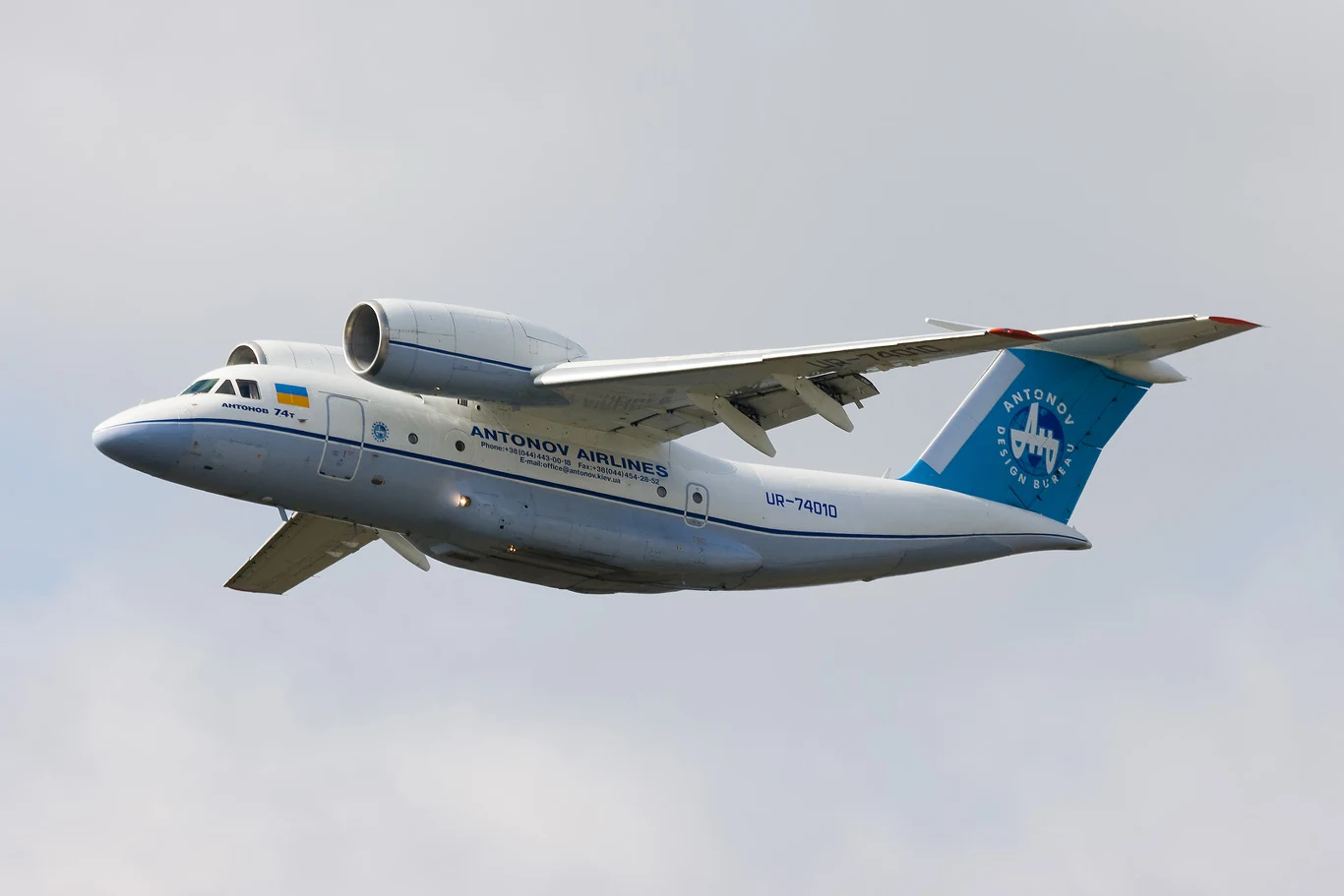 Duży samolot transportowy Antonov z niebieskim ogonem i napisem Antonov Airlines na kadłubie w locie na tle jasnego nieba.