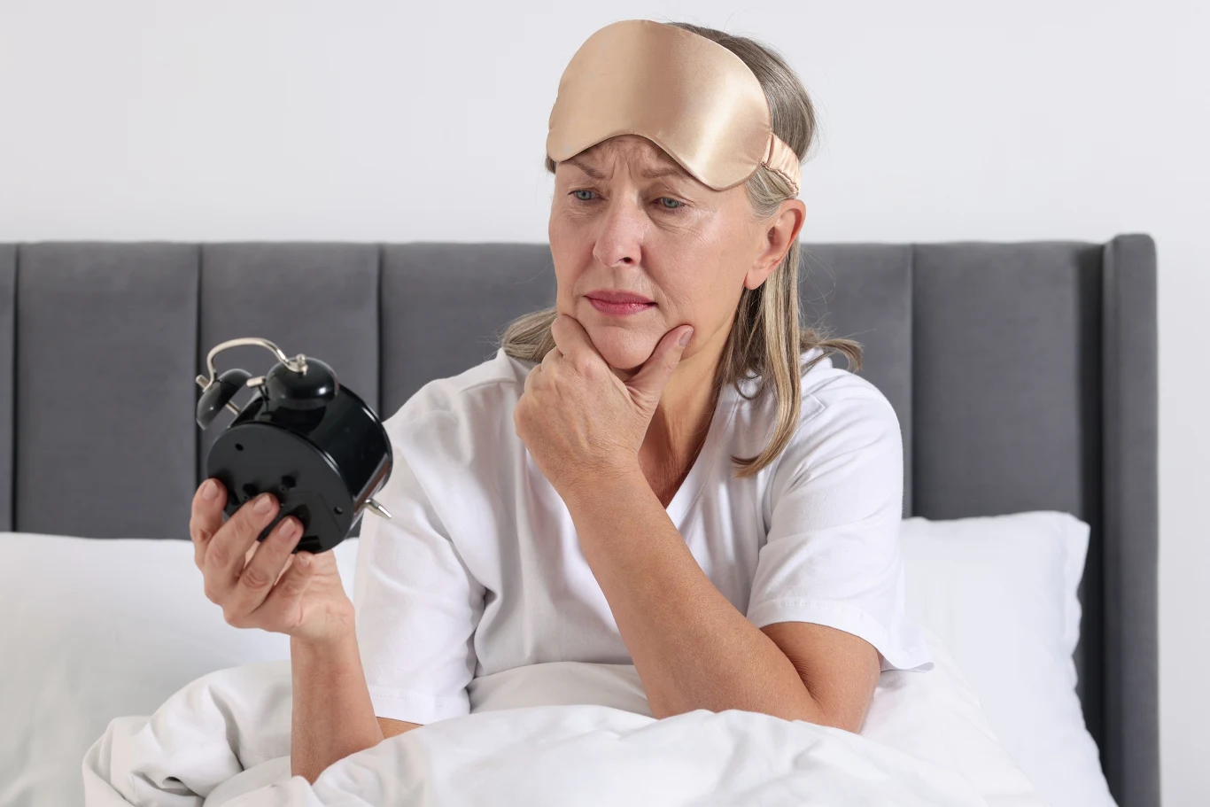 Starsza kobieta siedzi w łóżku w białej piżamie, z opaską na oczy zsuniętą na czoło, trzyma w ręku duży budzik i patrzy na niego z zamyśleniem.
