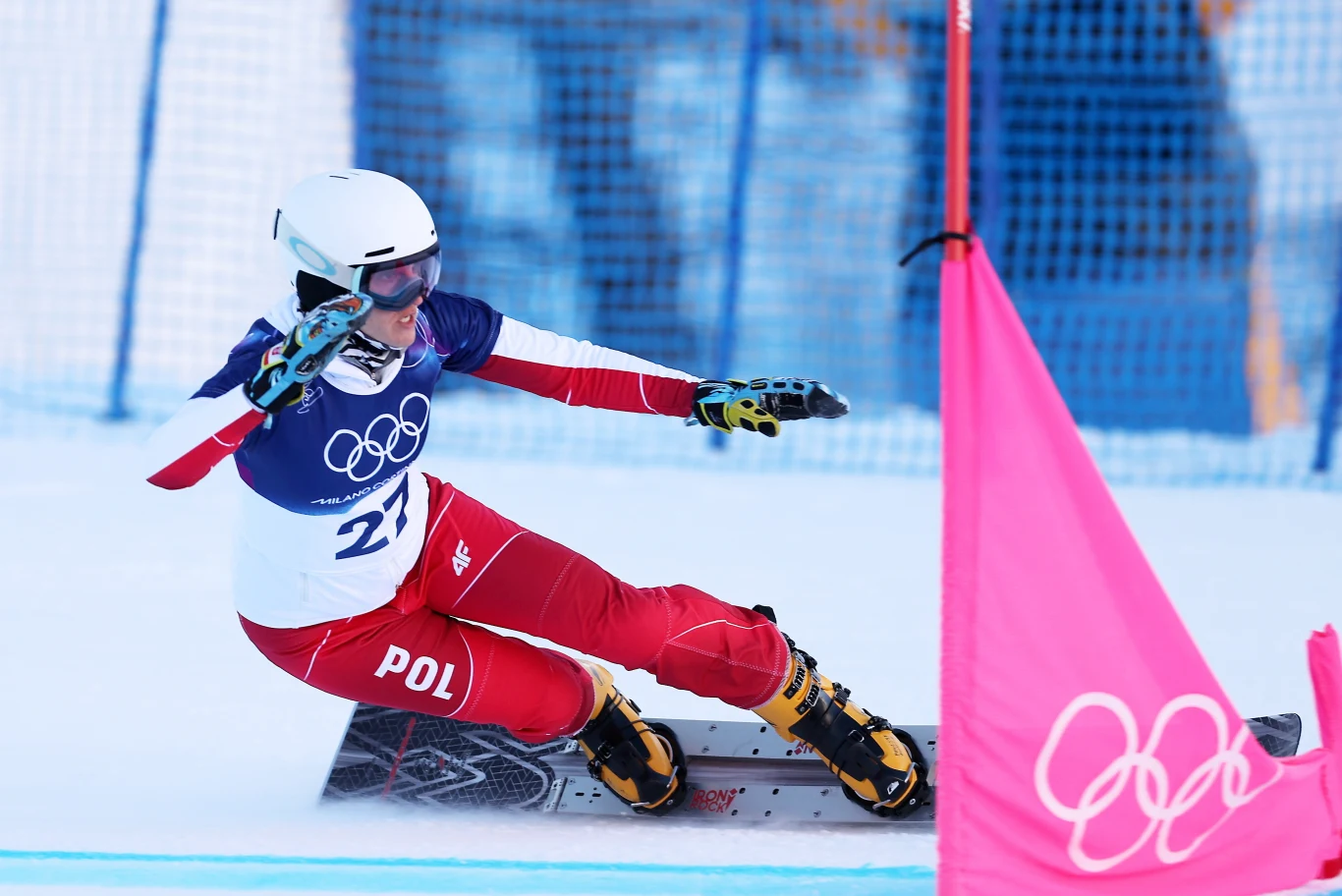 Aleksandra Król-Walas Zawodniczka na snowboardzie podczas wyścigu olimpijskiego, ubrana w biało-czerwony strój z numerem 27 i polską flagą. Przejeżdża dynamicznie obok różowego pachołka z logo igrzysk olimpijskich.
