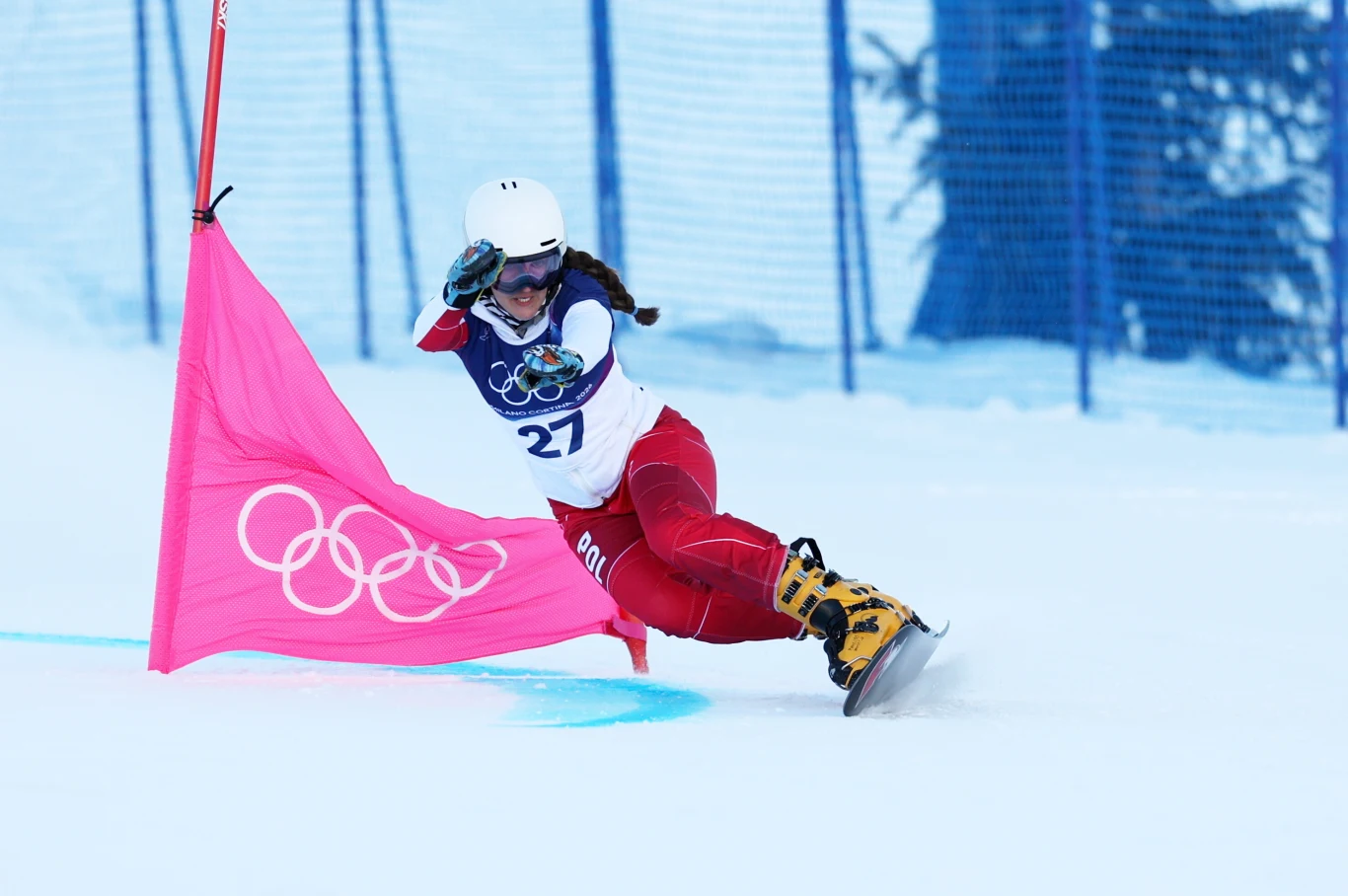 Aleksandra Król-Walas Zawodniczka w białym kasku i czerwonym stroju z numerem 27 wykonuje dynamiczny skręt podczas olimpijskiego wyścigu narciarskiego, mijając różową flagę z symbolem igrzysk olimpijskich na ośnieżonym stoku.
