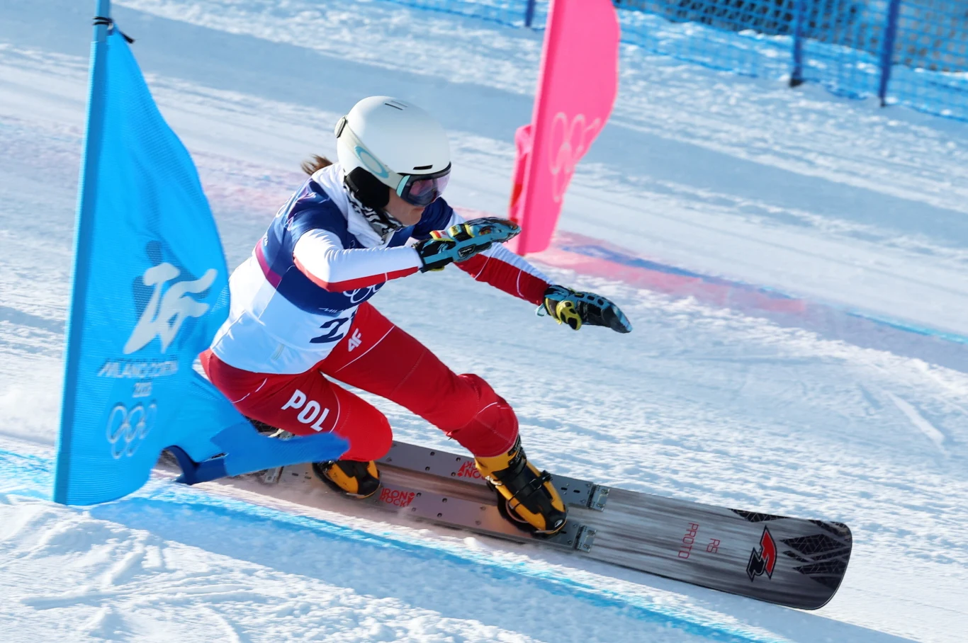 Aleksandra Król-Walas Narciarz snowboardowy w kasku i stroju reprezentacji Polski dynamicznie pokonuje slalom, przejeżdżając pomiędzy kolorowymi tyczkami na ośnieżonym stoku. W tle widoczne niebieskie ogrodzenie ochronne.