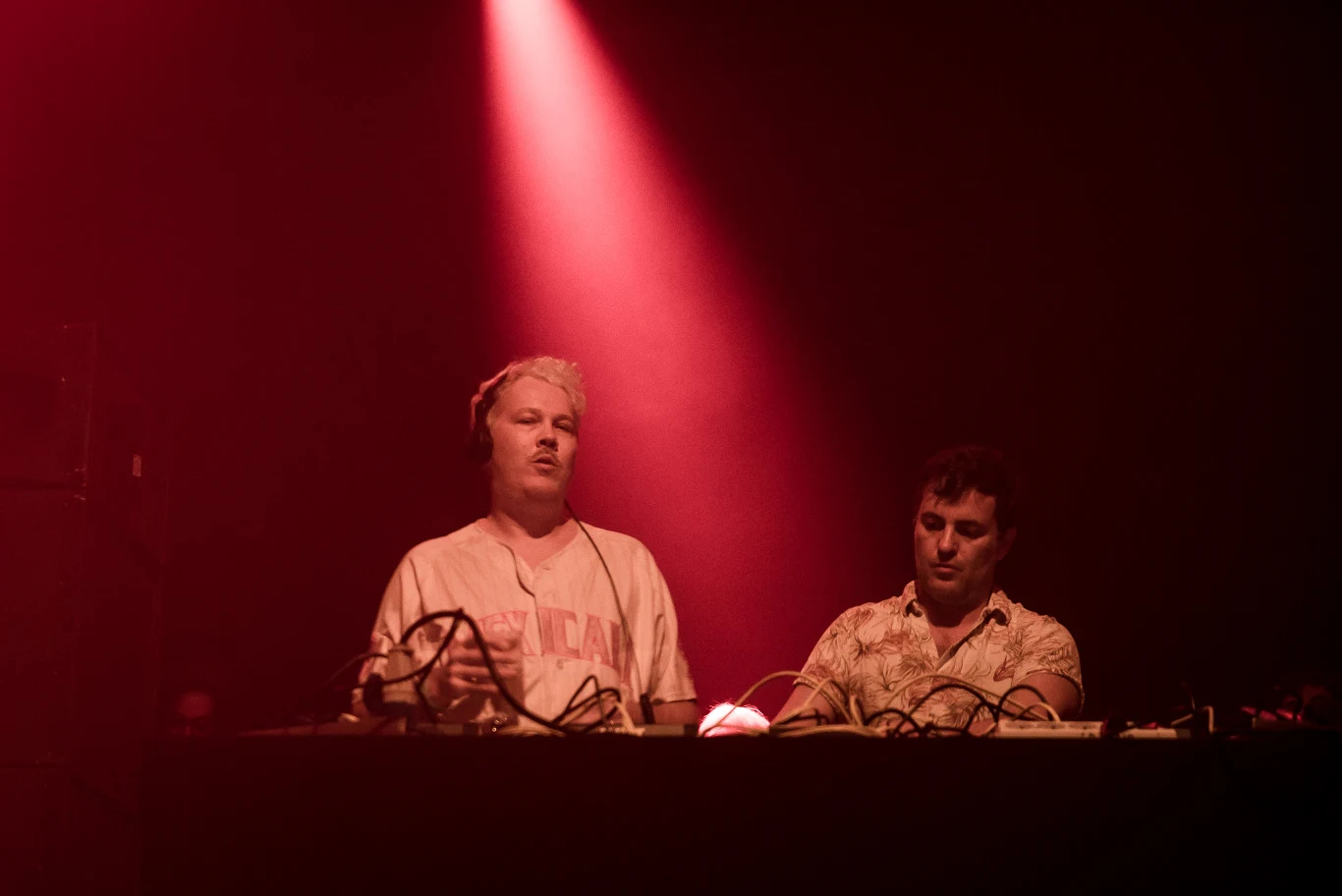 Dwóch DJ-ów stoi za konsoletą podświetloną czerwonym światłem, obsługując sprzęt muzyczny podczas występu na scenie.