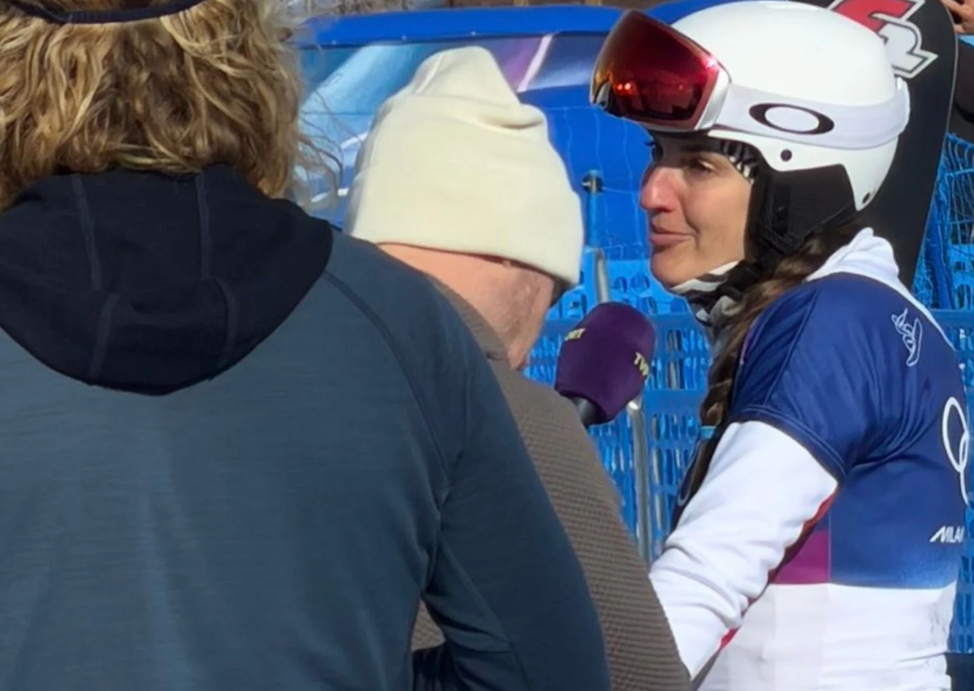 Aleksandra Król-Walas w rozmowie z mediami na igrzyskach olimpijskich Narciarka w stroju sportowym i kasku z goglami udziela wywiadu po zawodach. Osoba z mikrofonem ubrana w białą czapkę i kurtkę przysłania częściowo sportowca. W tle niebieskie ogrodzenia i śnieżna sceneria.