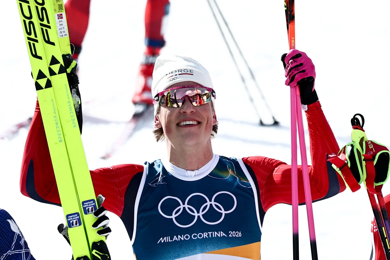 Zawodnik w sportowym stroju zimowym z logo igrzysk olimpijskich Milano Cortina 2026 unosi narty w geście triumfu i uśmiecha się szeroko. Ma na sobie okulary przeciwsłoneczne i białą czapkę, a w tle widoczne są rozmazane postacie innych narciarzy.