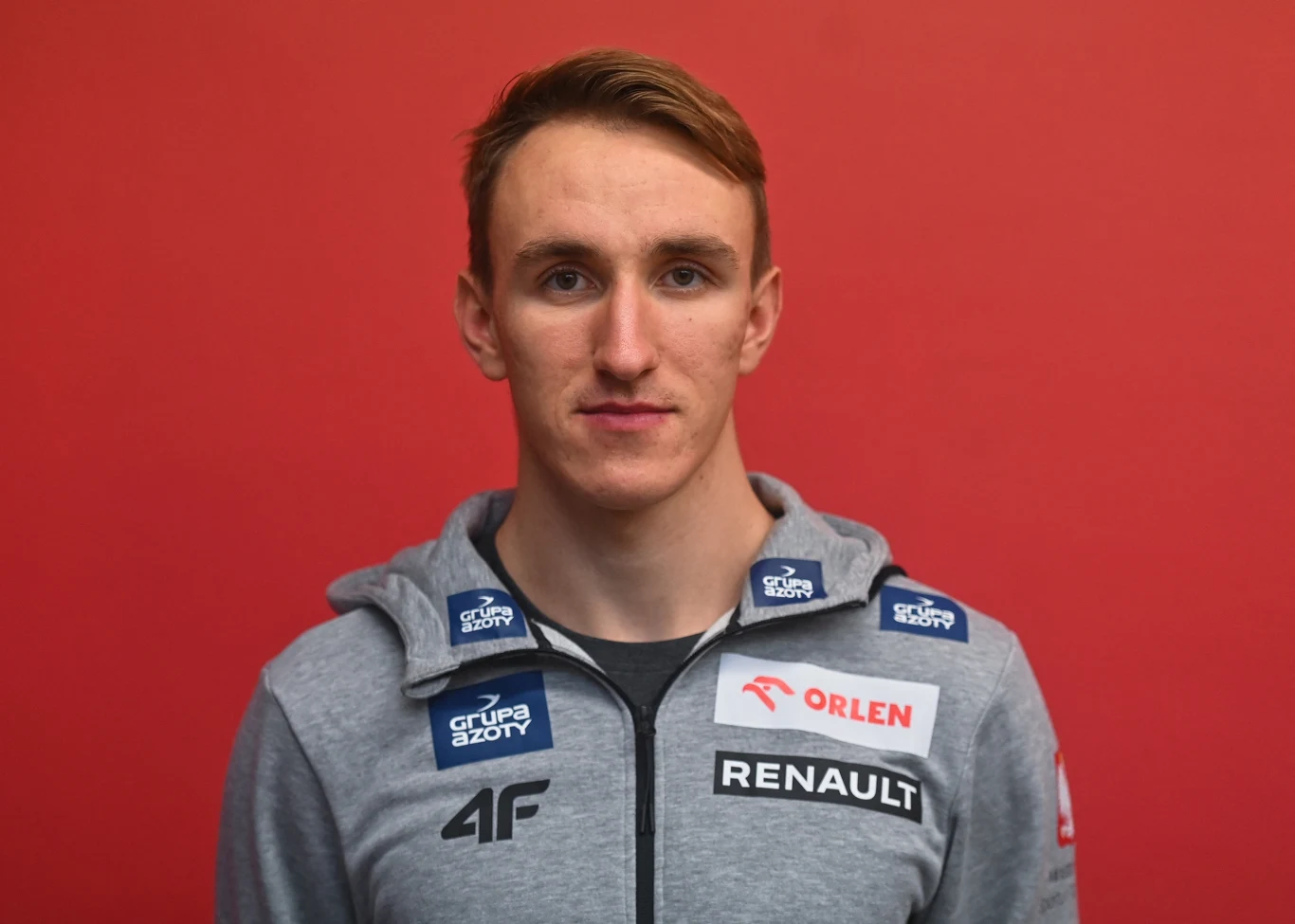 Młody mężczyzna w szarej bluzie sportowej z logotypami sponsorów na tle jednolitej, czerwonej ściany.