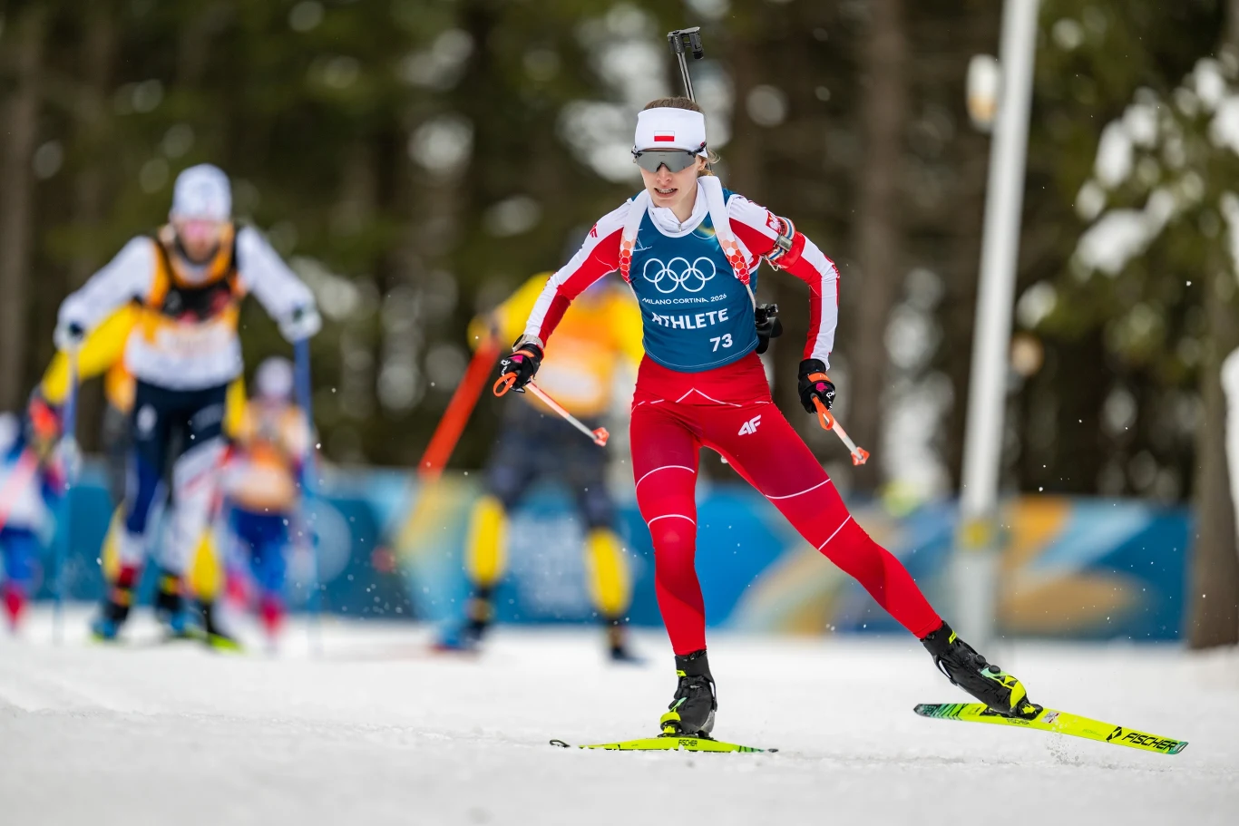 Natalia Sidorowicz Zawodniczka w czerwonym stroju z numerem 73 startująca w konkurencji biathlonowej podczas zimowych igrzysk olimpijskich, z karabinem na plecach. W tle inni zawodnicy na trasie, rozmyte drzewa i elementy scenografii olimpijskiej.