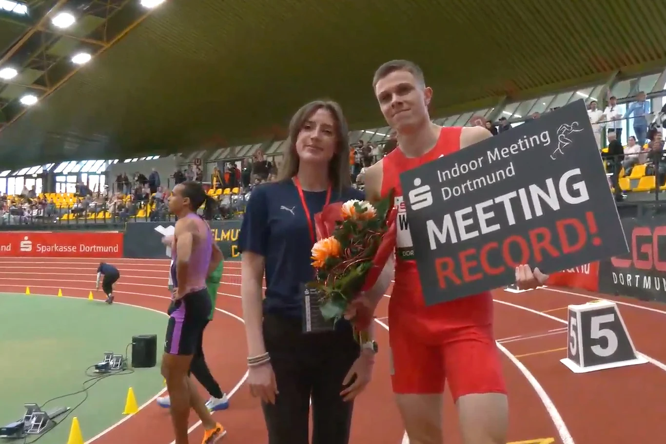 Lekkoatleta w czerwonym stroju trzyma tablicę z napisem 'MEETING RECORD!' oraz młodą kobietę z bukietem kwiatów. W tle hala sportowa oraz inni uczestnicy zawodów lekkoatletycznych.