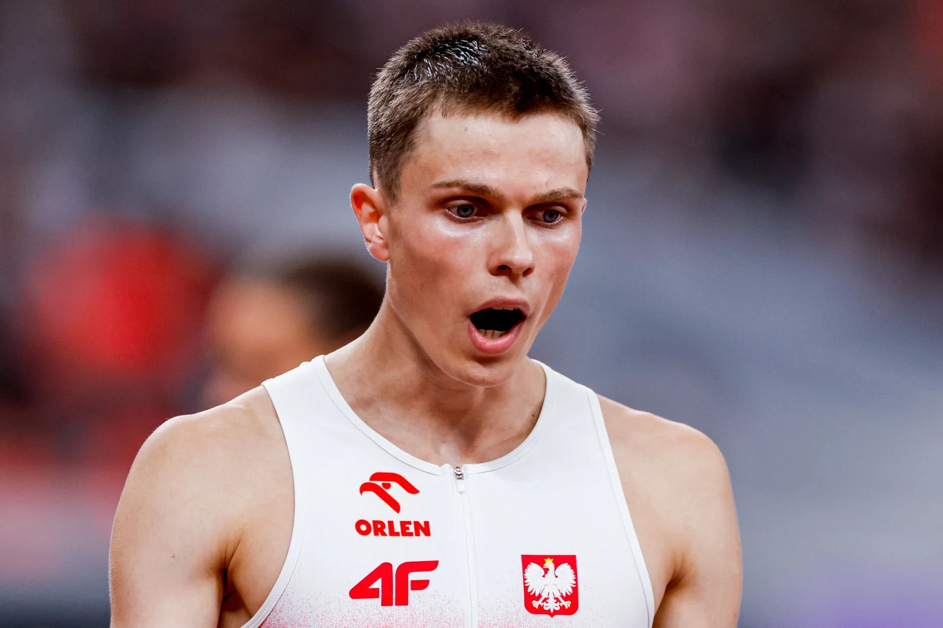 Młody lekkoatleta ubrany w biały strój sportowy z wyraźnymi emblematami Polski i sponsora, z otwartymi ustami, prawdopodobnie w reakcji na emocje po wykonanym biegu, w tle rozmyta publiczność stadionu.
