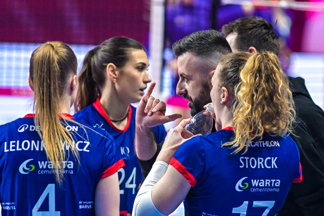 Trener udziela wskazówek drużynie siatkówki podczas krótkiej przerwy w meczu, zawodniczki uważnie słuchają, koncentrując się na strategii gry, widoczne sportowe stroje z nazwiskami i logotypami sponsorów.