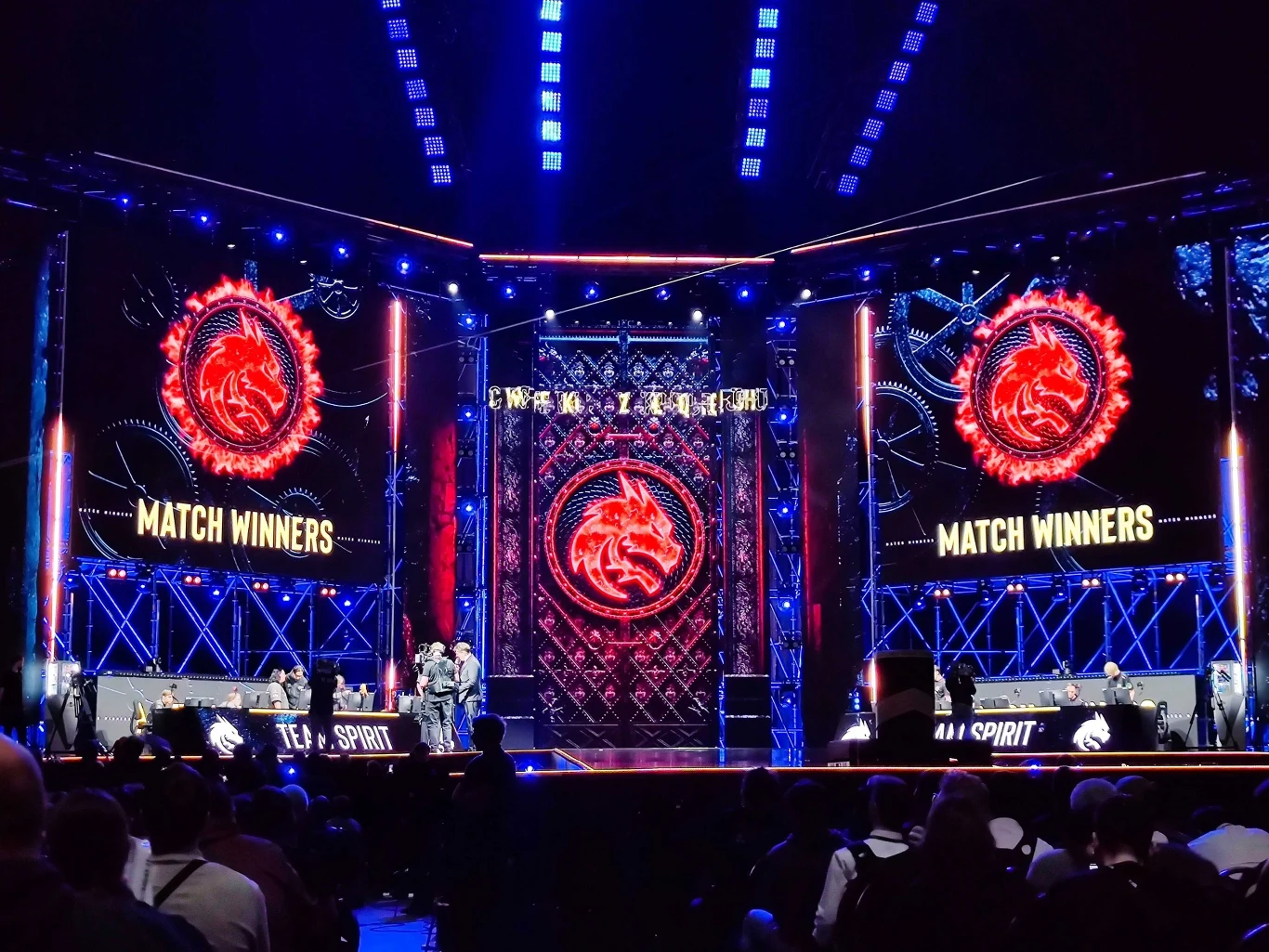 Duża scena turniejowa podczas wydarzenia e-sportowego, na której na ekranach widoczny jest napis 'MATCH WINNERS' oraz logo drużyny Team Spirit. Po bokach sceny siedzą zawodnicy przy stanowiskach komputerowych, publiczność obserwuje całość z widowni.