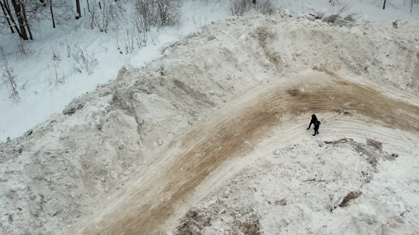 Osoba ubrana na ciemno idzie szeroką, zakręcającą, ubitą ścieżką śnieżną pośrodku dużej zaspy w zimowym krajobrazie, wokół śnieg i drzewa.