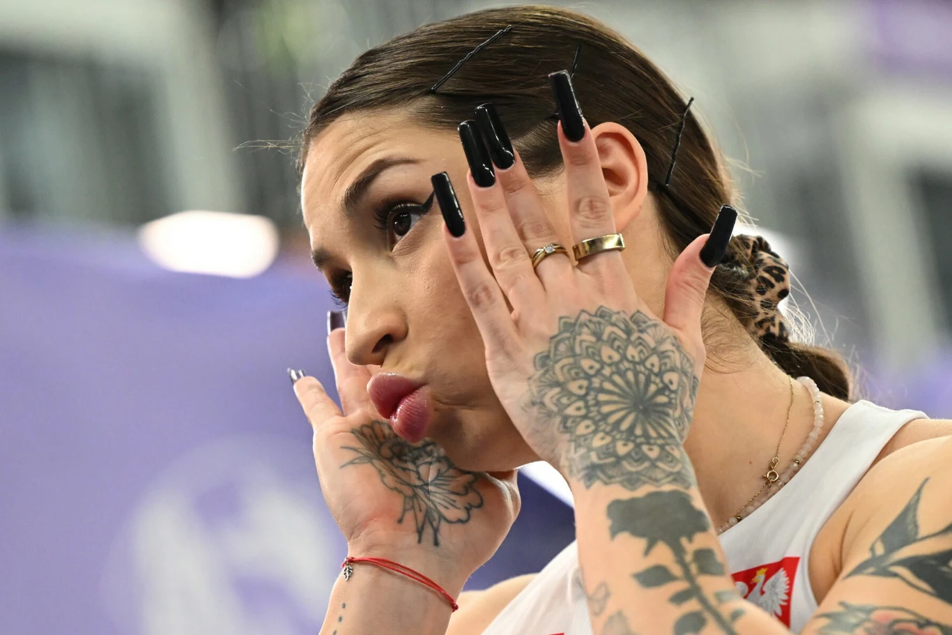 Młoda kobieta z długimi, czarnymi tipsami pozuje z dłońmi przy twarzy, eksponując liczne tatuaże na rękach, twarz w wyrazie skupienia lub zabawy, sportowy strój z polskim godłem widoczny na ramieniu.