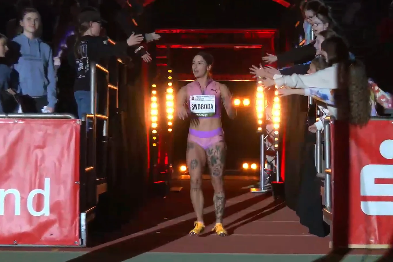 Lekkoatletka ubrana w strój sportowy wchodzi na arenę startową, po bokach ustawieni kibice wyciągają ręce w jej stronę, w tle widać mocne światła oświetlające przejście.