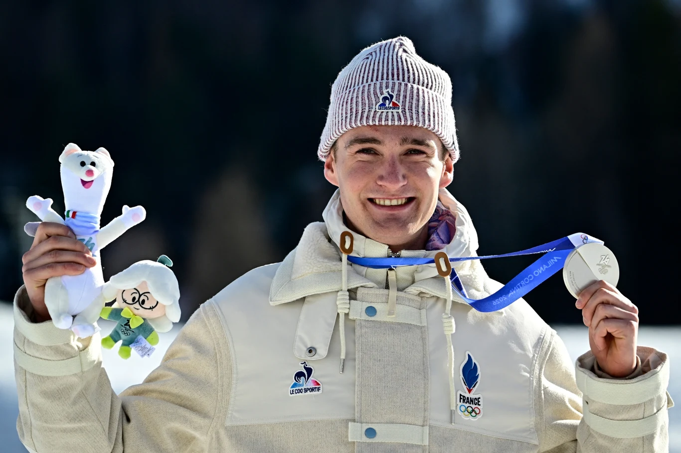 Uśmiechnięty młody mężczyzna w jasnej kurtce i czapce z logo reprezentacji Francji trzyma w jednej dłoni maskotkę, a w drugiej srebrny medal na niebieskiej wstążce. W tle rozmyte drzewa i zimowa sceneria.
