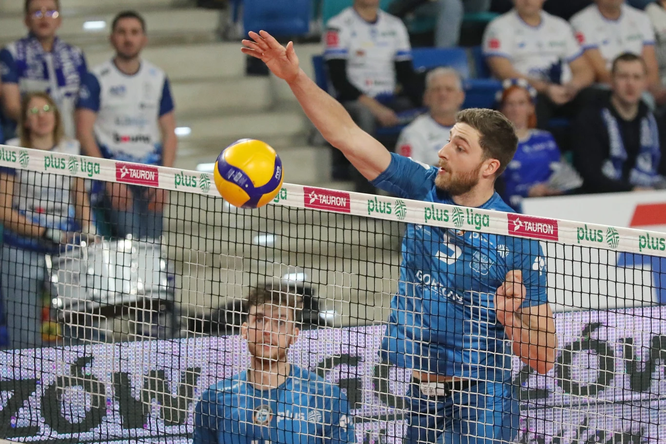 Zawodnik drużyny siatkarskiej ubrany w niebieski strój wykonuje atak nad siatką, piłka zmierza w kierunku przeciwnika, w tle widoczna publiczność oraz inni gracze.