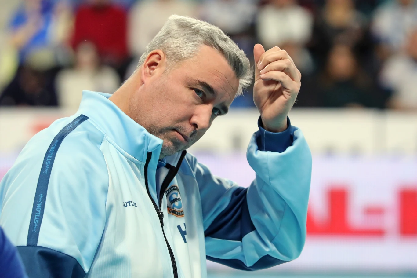 Mężczyzna w sportowej kurtce w jasnoniebieskich odcieniach stoi na pierwszym planie, wykonując gest ręką przy czole, w tle rozmyta publiczność lub widownia.