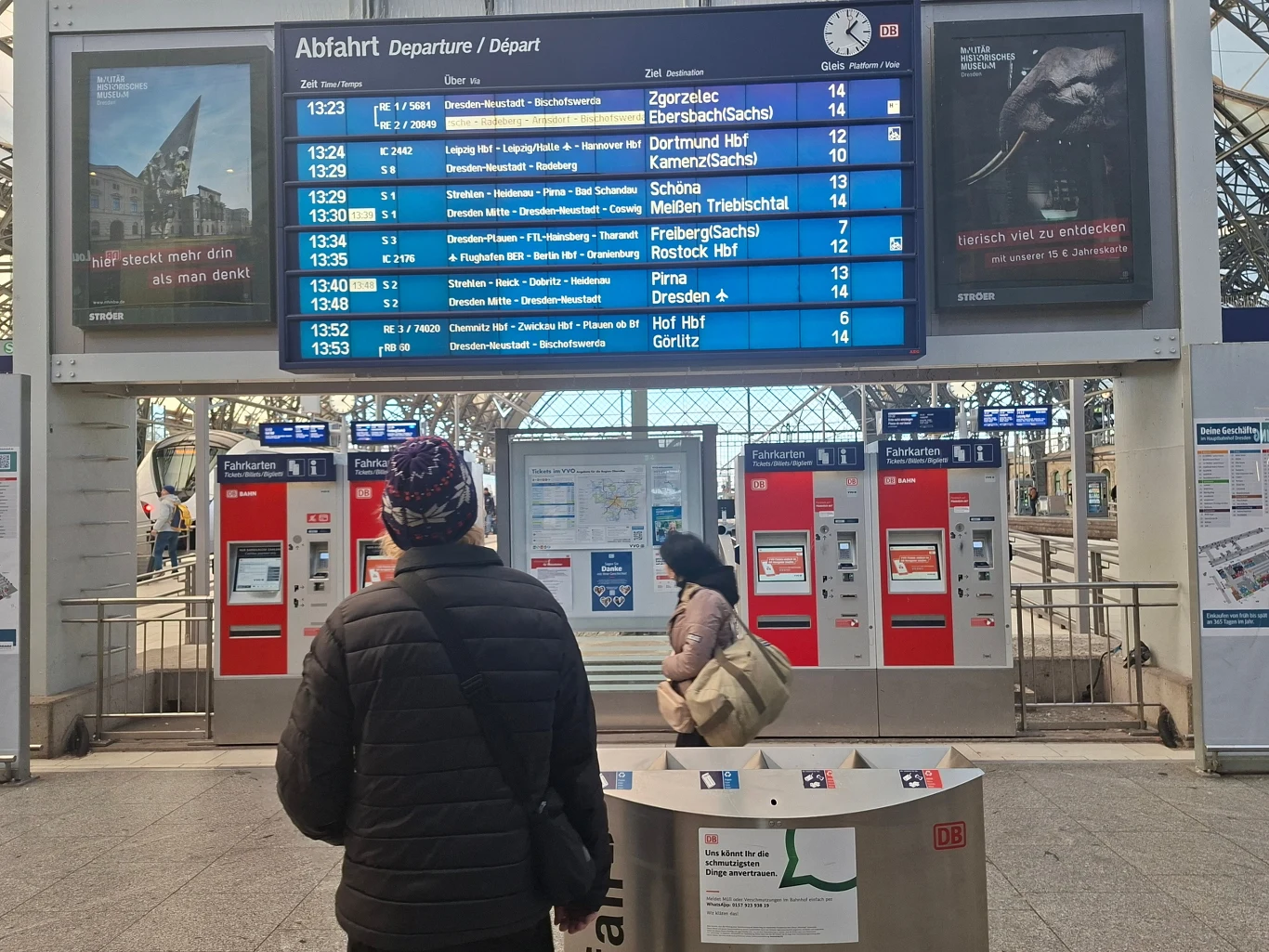 Duża elektroniczna tablica odjazdów z informacjami o pociągach, automatami biletowymi poniżej oraz dwie osoby patrzące na rozkład jazdy w hali dworca kolejowego.