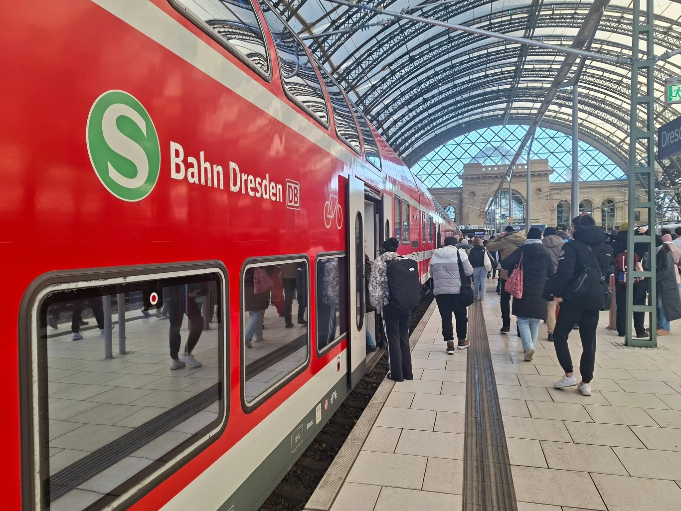 Dwupiętrowy czerwony pociąg z napisem S-Bahn Dresden stoi przy peronie w zadaszonej hali dworca, obok pociągu stoi i wsiada grupa osób z plecakami i torbami, widoczna nowoczesna architektura ze stalowym szkieletem i szklaną ścianą.