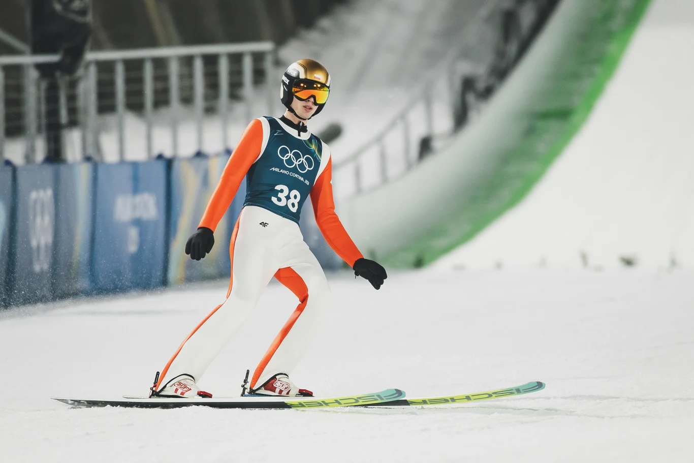 Kacper Tomasiak Zawodnik w kombinezonie narciarskim ląduje na nartach podczas zawodów olimpijskich w skokach narciarskich, w tle widoczny fragment skoczni oraz barierki zabezpieczające.