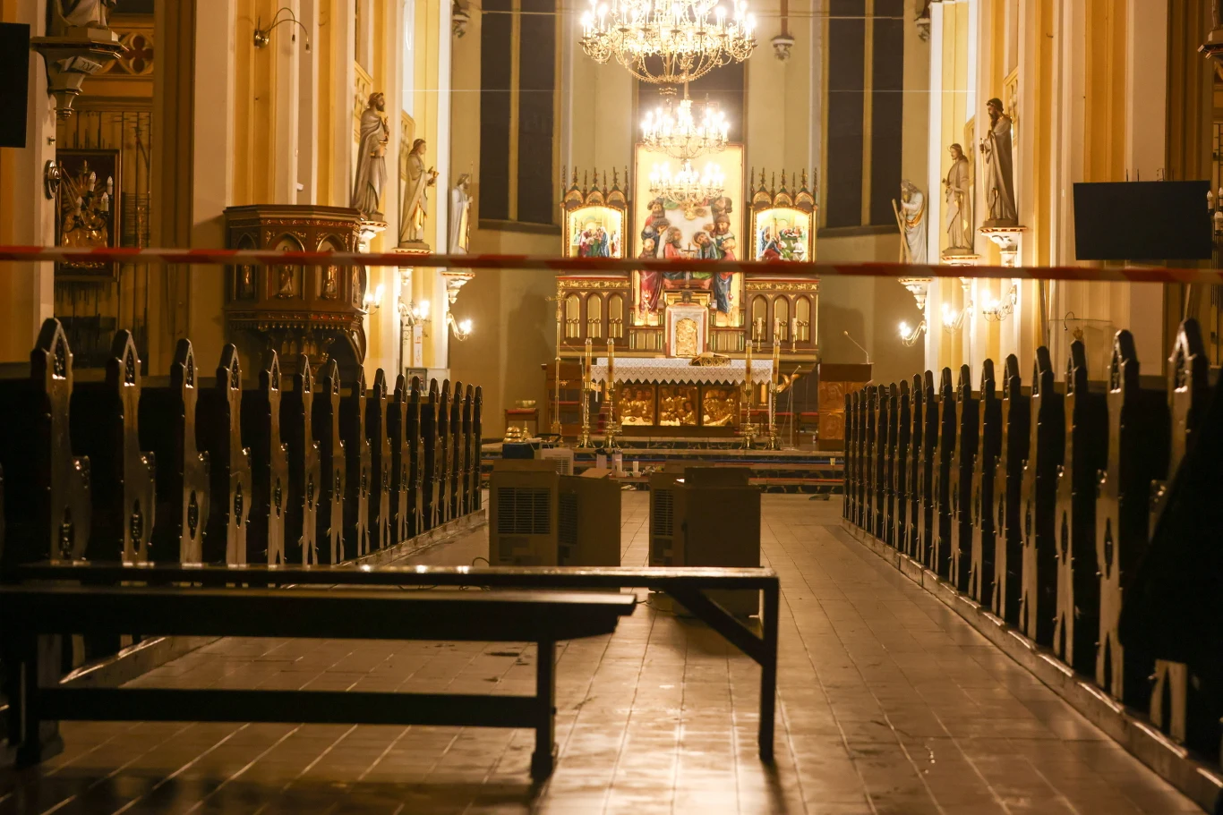 Wnętrze kościoła z rzędem ław po obu stronach nawy, ołtarz z bogato zdobionymi ikonami i figurami, jasne światło żyrandoli podkreśla detale złotych dekoracji, a przód oddzielony jest taśmą.