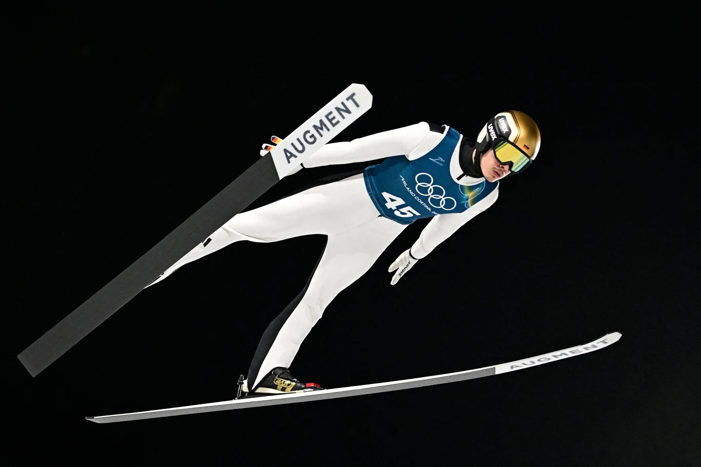 Philipp Raimund Skoczek narciarski w białym kombinezonie podczas lotu w powietrzu z numerem startowym 45, złotym kaskiem oraz nartą z widocznym logo AUGMENT na tle czarnego nieba.