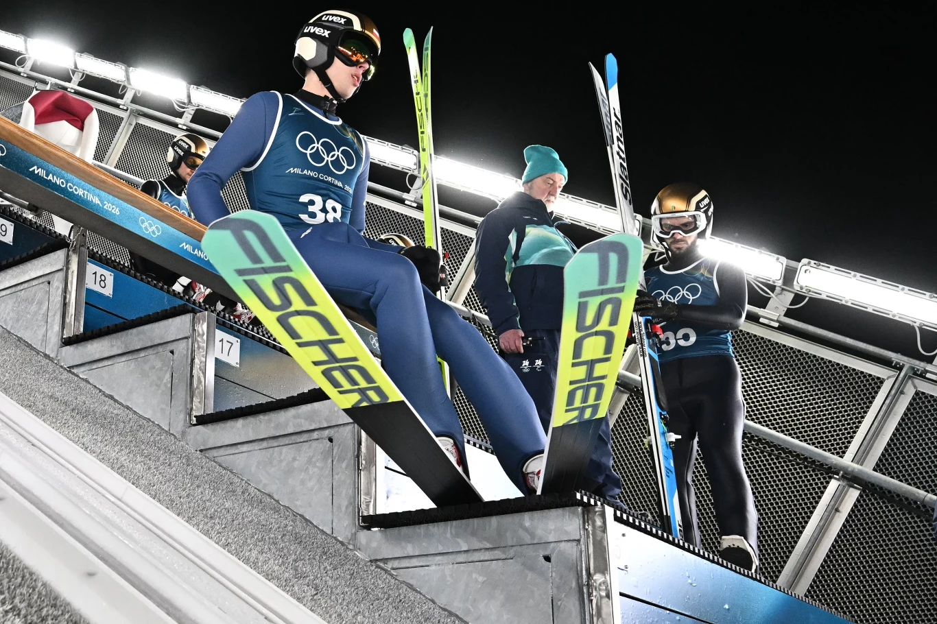 Zawodnik w kombinezonie narciarskim z numerem 38 przygotowuje się do skoku narciarskiego, siedząc na belce startowej na dużej skoczni, obok widać innych skoczków oraz trenerów w sportowych ubraniach, na twarzach skupienie, w tle nocne oświetlenie.