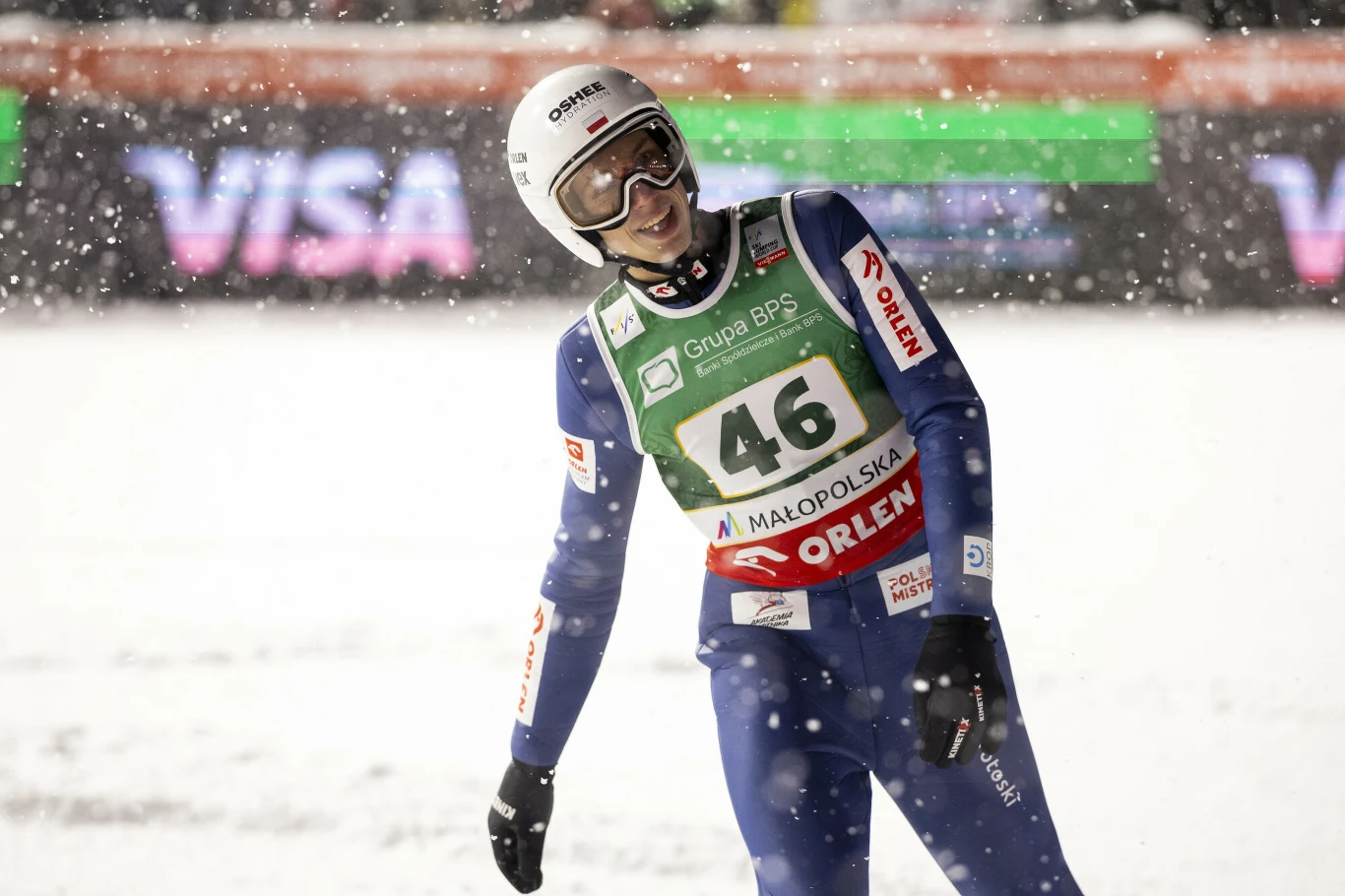 Zawodnik w kombinezonie narciarskim i kasku stoi na śnieżnej skoczni podczas opadów śniegu. Widoczne są plakietki sponsorów oraz numer startowy 46. W tle rozmazane światła i banery reklamowe.