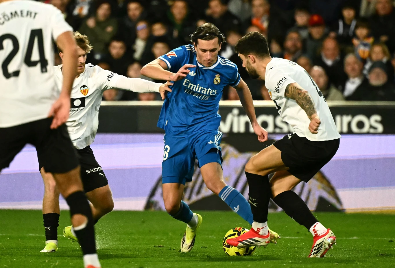 Piłkarz w niebieskim stroju drybluje z piłką pomiędzy dwoma obrońcami w białych koszulkach podczas meczu piłkarskiego, wokół zawodników znajduje się tłum kibiców na trybunach.