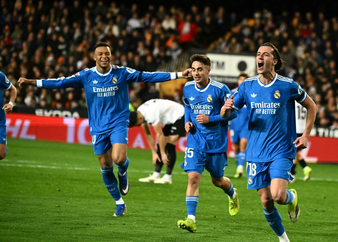 Zawodnicy drużyny Real Madryt w niebieskich strojach cieszą się po zdobyciu gola podczas meczu piłkarskiego, w tle widoczni są zawodnicy przeciwnej drużyny oraz kibice na trybunach.