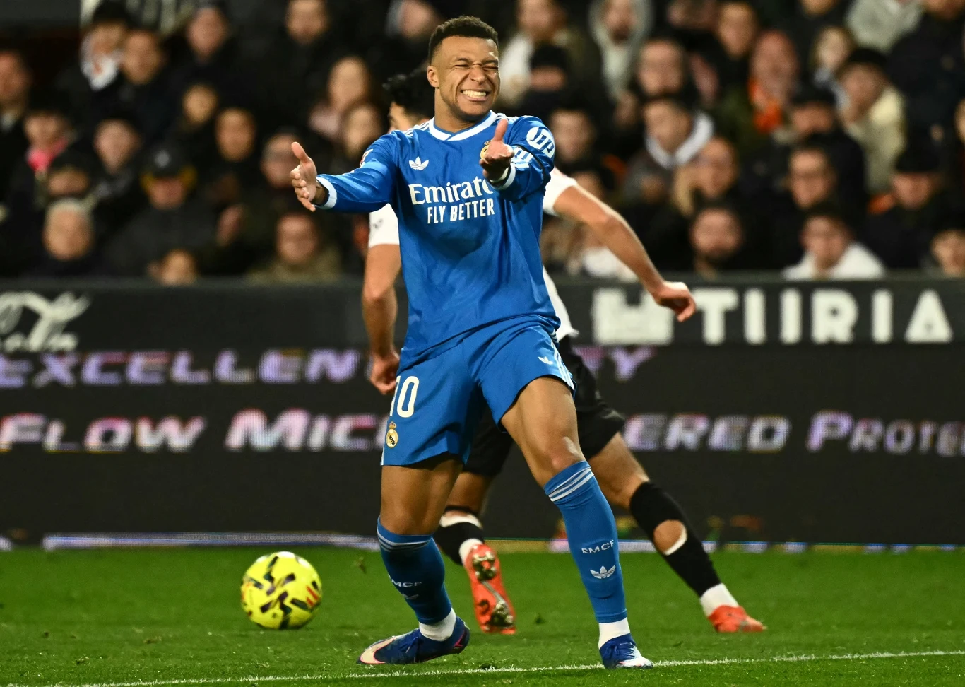 Piłkarz w niebieskim stroju Realu Madryt wykonuje dynamiczny gest podczas meczu piłkarskiego, wokół znajdują się inni zawodnicy oraz widzowie na trybunach, na pierwszym planie znajduje się piłka.