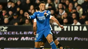 Piłkarz w niebieskim stroju Realu Madryt wykonuje dynamiczny gest podczas meczu piłkarskiego, wokół znajdują się inni zawodnicy oraz widzowie na trybunach, na pierwszym planie znajduje się piłka.