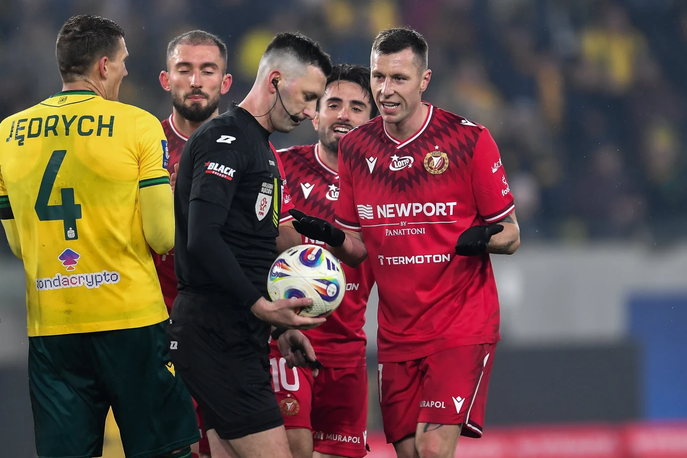 Grupa piłkarzy wraz z sędzią na boisku podczas meczu piłki nożnej, zawodnik w czerwonym stroju gestykuluje w kierunku arbitra trzymającego piłkę, obok stoją zawodnicy w żółto-zielonych koszulkach.