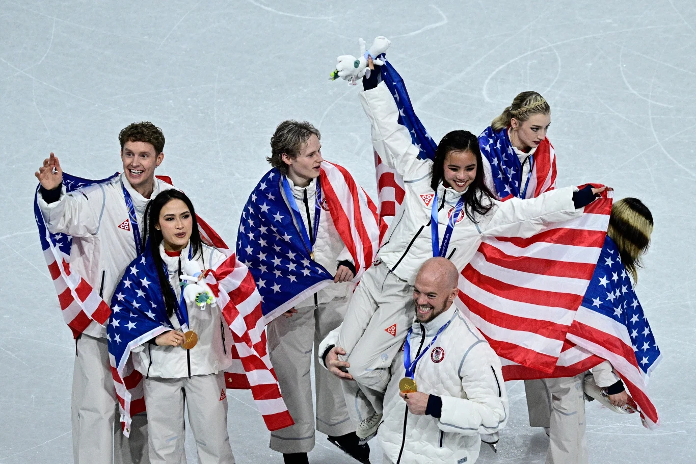 Grupa sportowców ubranych w białe stroje trzyma amerykańskie flagi, wszyscy mają na szyjach medale. Uśmiechają się, celebrując sukces na lodowej arenie.