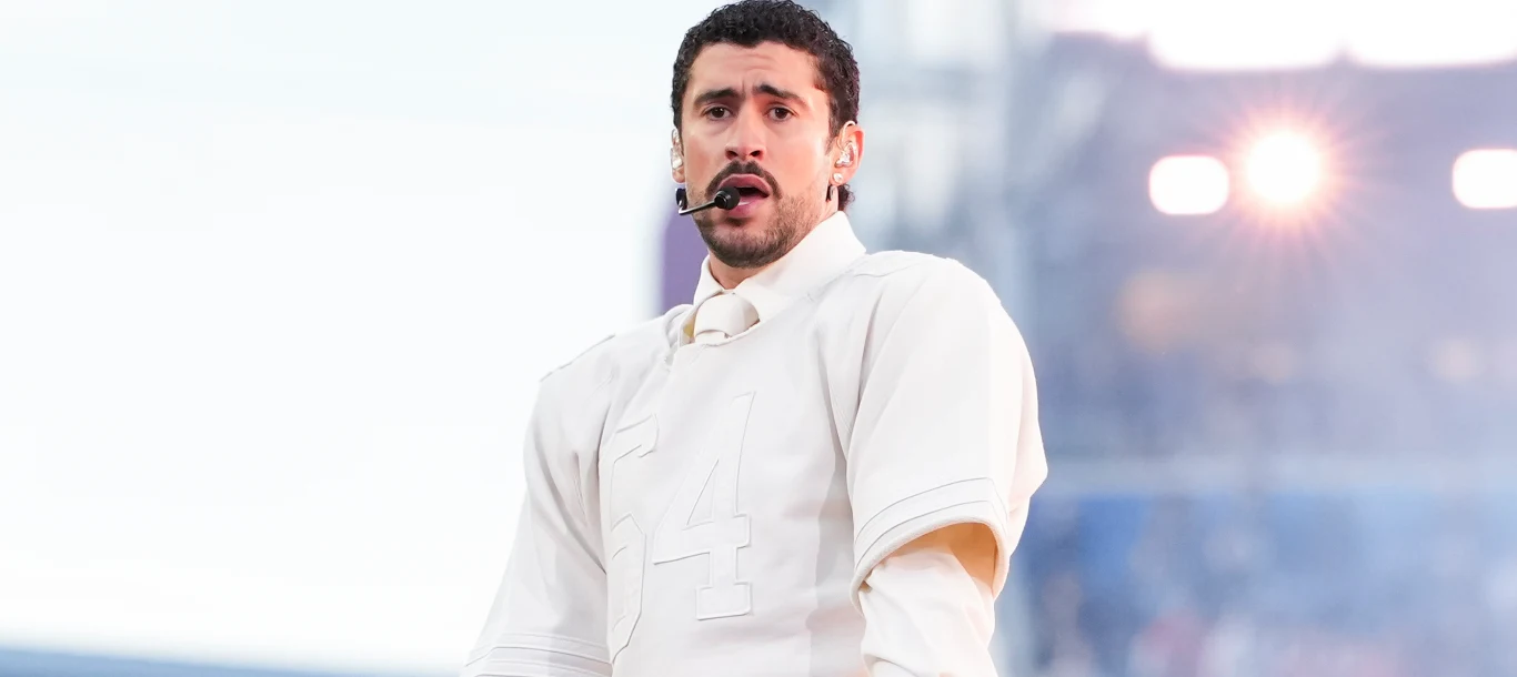 Bad Bunny Mężczyzna z krótkimi, ciemnymi włosami i zarostem, ubrany w białą koszulę oraz kamizelkę z numerem 4, ze słuchawką mikrofonową na głowie, stoi na tle rozświetlonego światła scenicznego.