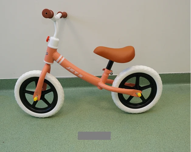 Rower biegowy dziecięcy w kolorze pomarańczowym z białymi oponami i czarnymi felgami stoi na zielonej podłodze przy ścianie.