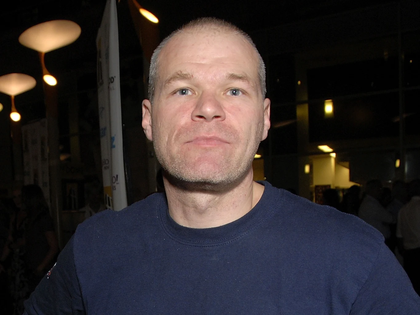 Uwe Boll w 2007 roku Mężczyzna o krótkich, jasnych włosach i jasnej cerze patrzy prosto w obiektyw, ubrany w granatową koszulkę, w tle widoczne są rozmyte światła oraz sylwetki innych osób.