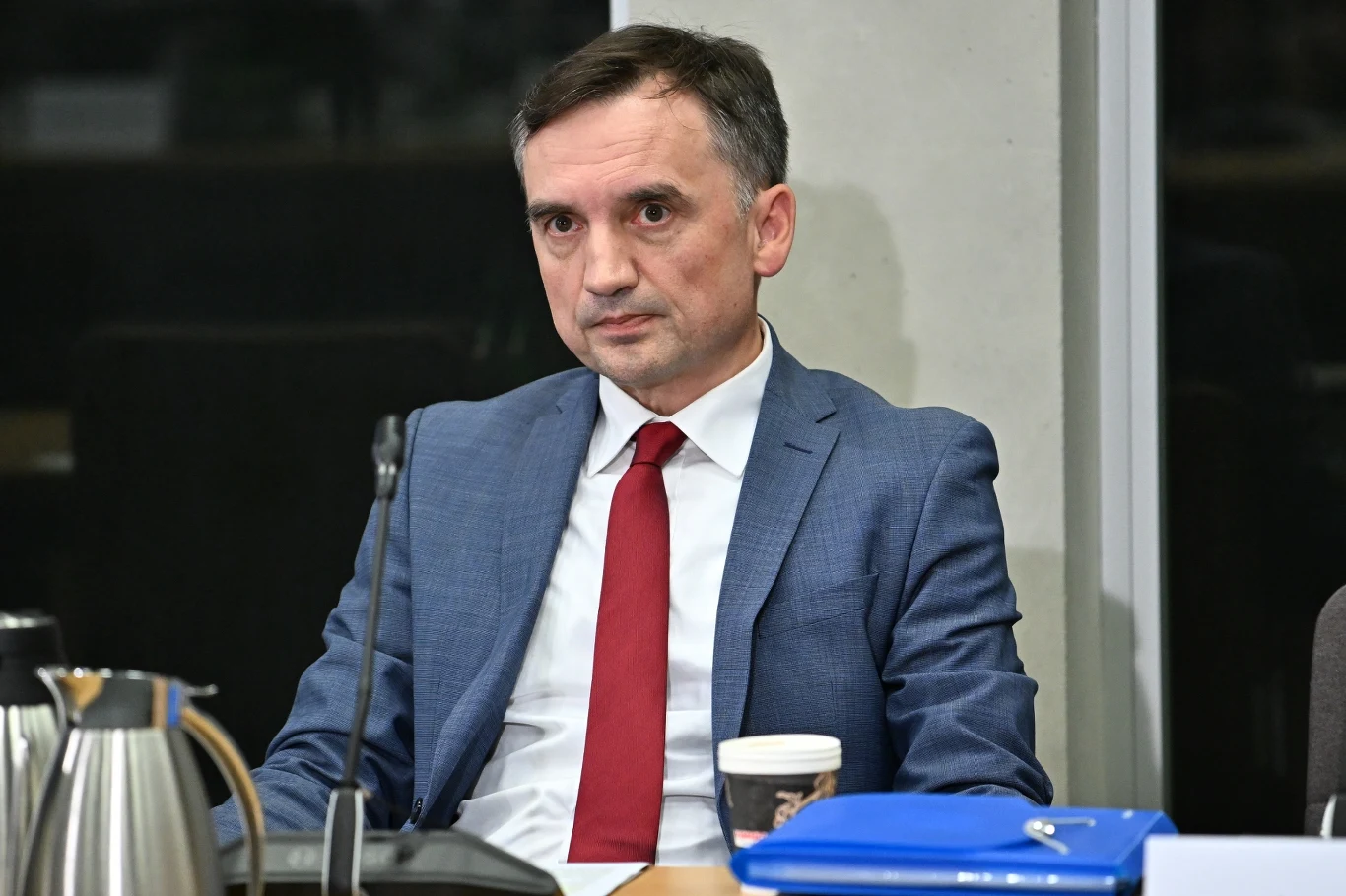 Zbigniew Ziobro Mężczyzna w garniturze z czerwoną krawatą siedzi przy stole konferencyjnym, przed nim leży niebieska teczka, filiżanka oraz dzbanek metalowy.